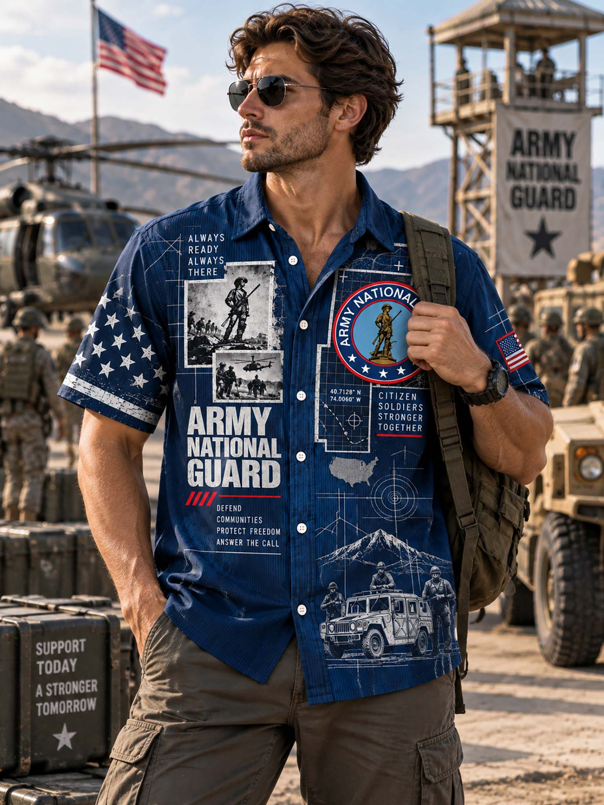 A man wearing a blue short-sleeve button-up shirt with Army National Guard graphics, standing in a military base setting with military vehicles and an American flag in the background.