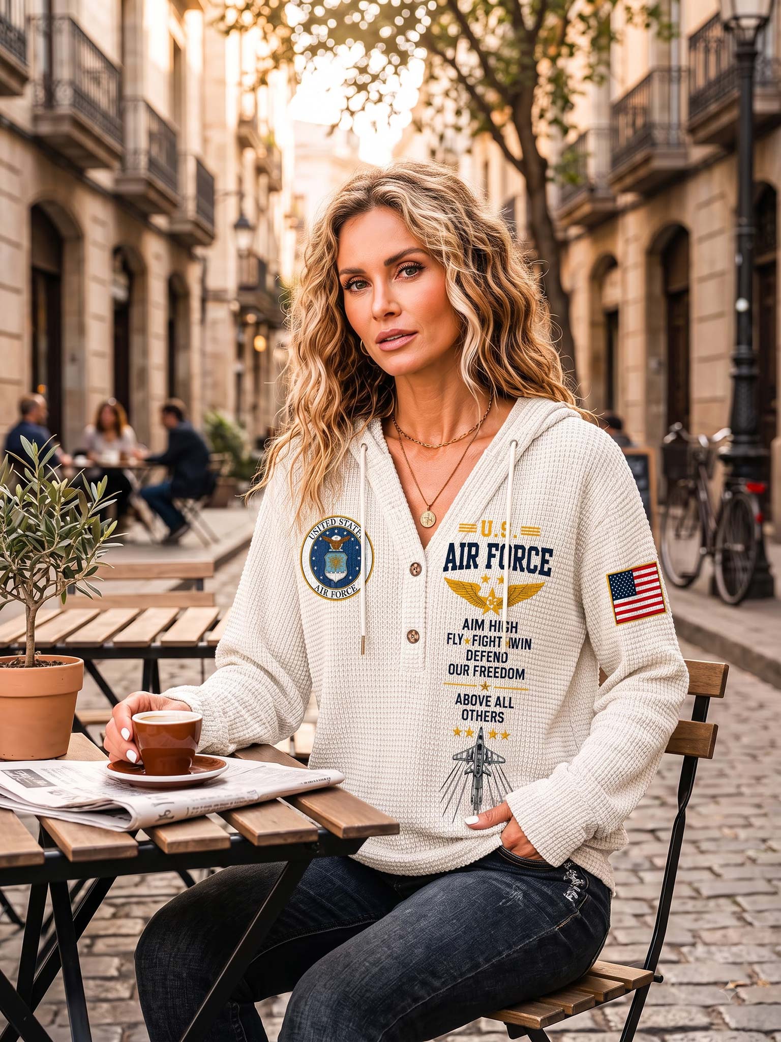 A woman sitting at an outdoor cafe table wearing a cream-colored waffle-knit hoodie with 'AIR FORCE' text and military insignia patches, paired with dark jeans.