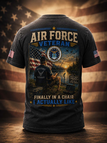 A dark charcoal gray t-shirt shown from the back, featuring a large graphic print with the text 'AIR FORCE VETERAN' and 'FINALLY IN A CHAIR I ACTUALLY LIKE'. The graphic depicts a military officer, an American flag, a lake at sunset, and a sign reading 'SOMETIMES I SIT ON A LOG ANCHOR AWE'.