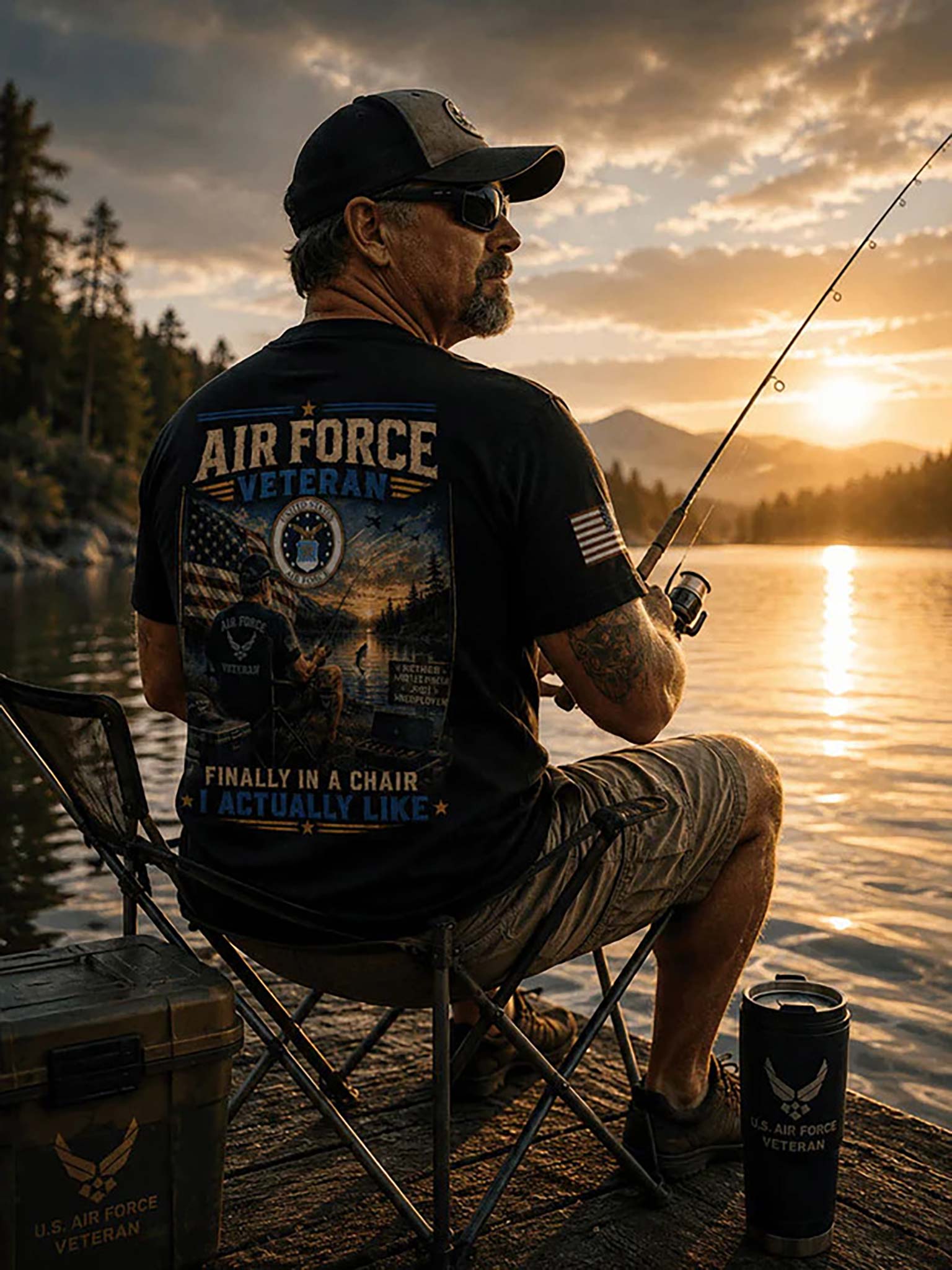 A man sits on a folding chair on a dock by a lake at sunset, wearing a black graphic T-shirt with 'AIR FORCE VETERAN' text and imagery on the back, khaki shorts, and a cap. A fishing rod extends from his hand, and a U.