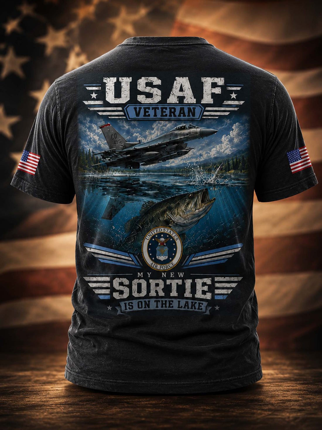 A black graphic t-shirt shown from the back, featuring a large design with a military jet flying over water, a fish, and the text 'USAF VETERAN' and 'MY NEW SORTIE IS ON THE LAKE'. The shirt also has small American flag prints on both sleeves, set against a blurred American flag background.