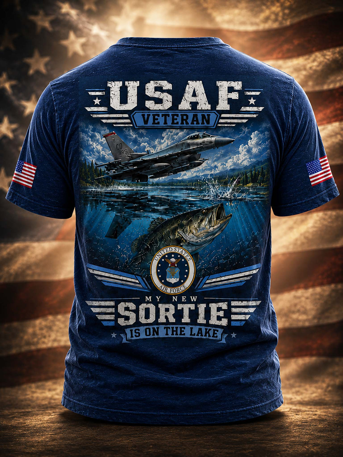A black graphic t-shirt shown from the back, featuring a large design with a military jet flying over water, a fish, and the text 'USAF VETERAN' and 'MY NEW SORTIE IS ON THE LAKE'. The shirt also has small American flag prints on both sleeves, set against a blurred American flag background.