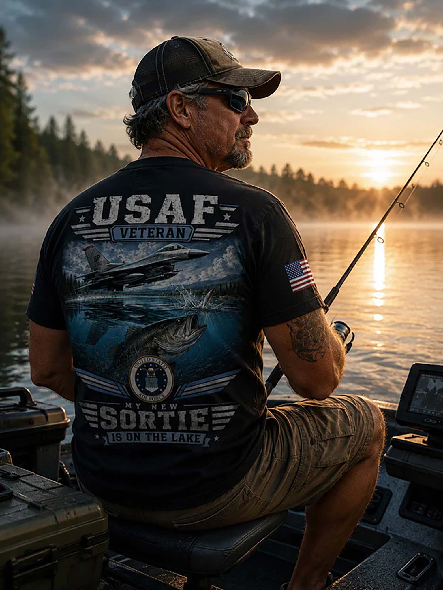 A man sitting in a boat on a lake at sunset, wearing a black t-shirt with a large blue and white USAF Veteran graphic on the back, khaki shorts, and a black cap.