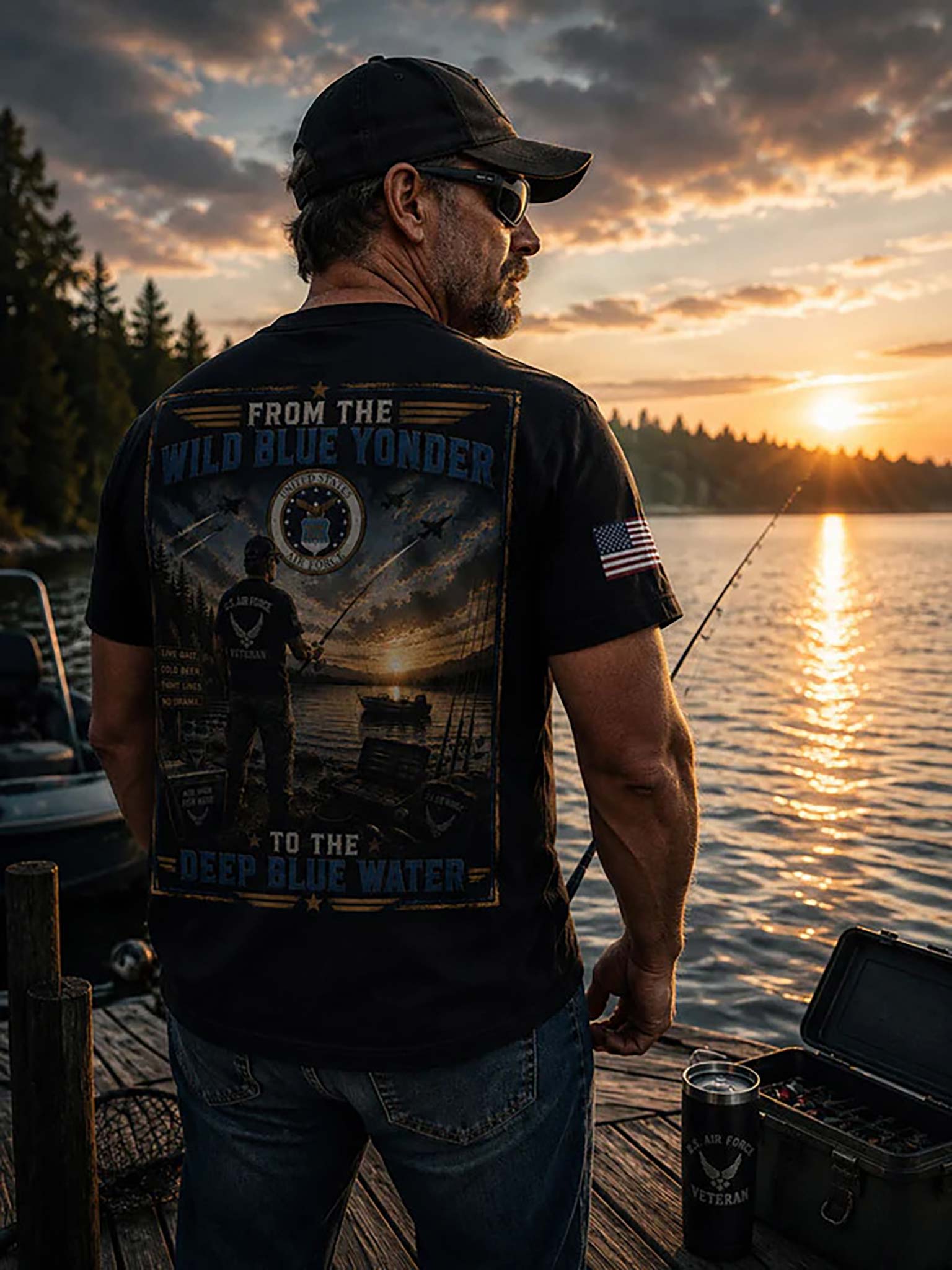 A man standing on a wooden dock at sunset wearing a black graphic t-shirt with a large back print reading 'FROM THE WILD BLUE YONDER TO THE DEEP BLUE WATER' and a small American flag patch on the sleeve, fishing by the water.
