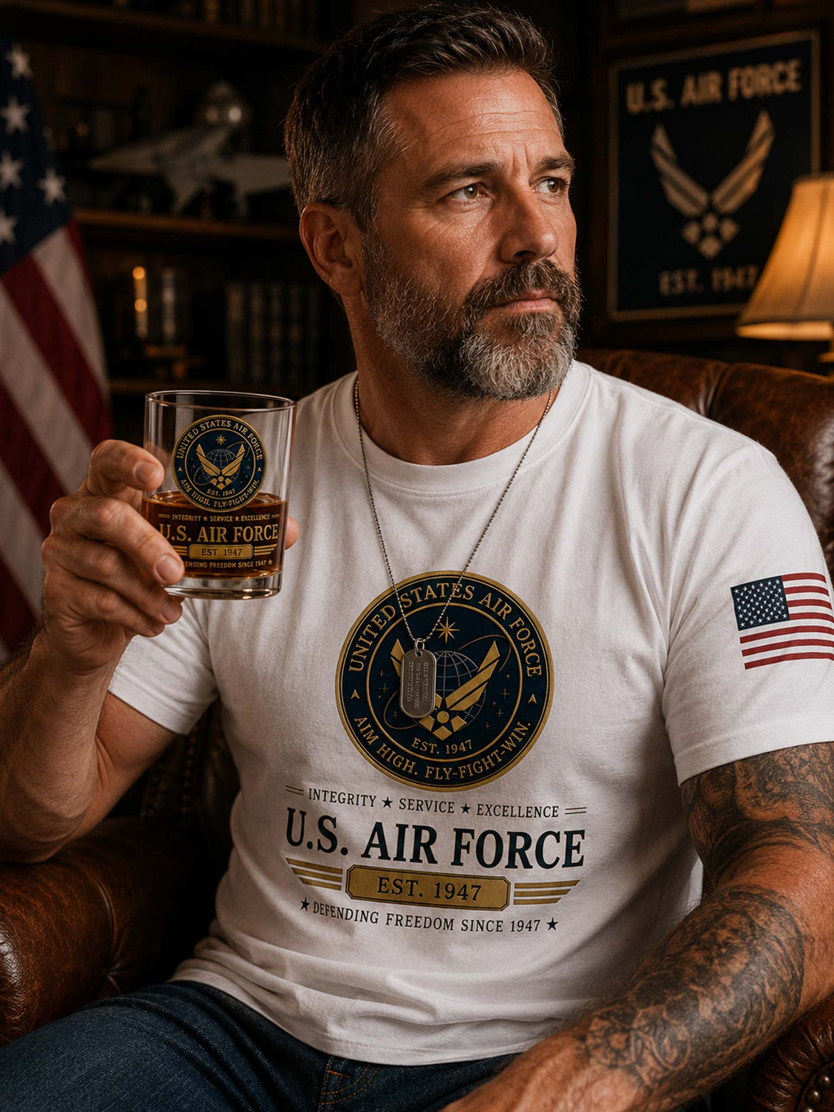 A man with a beard and tattoos sits in a leather armchair holding a glass of whiskey. He is wearing a white graphic t-shirt featuring a United States Air Force emblem on the chest and an American flag on the sleeve, with a U.