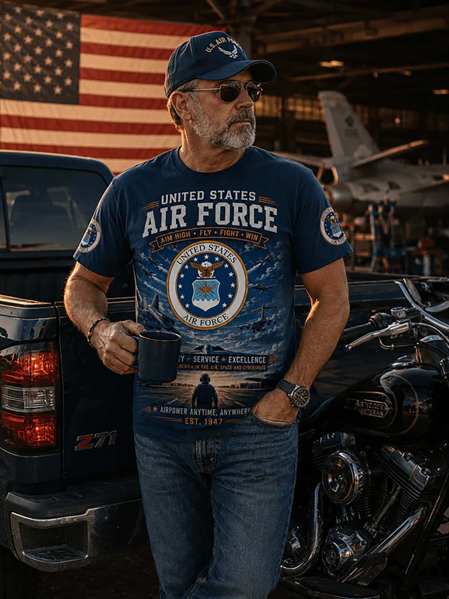 A man wearing a navy blue graphic t-shirt with 'United States Air Force' text and insignia, standing in front of a black pickup truck and motorcycle in a garage with an American flag in the background.