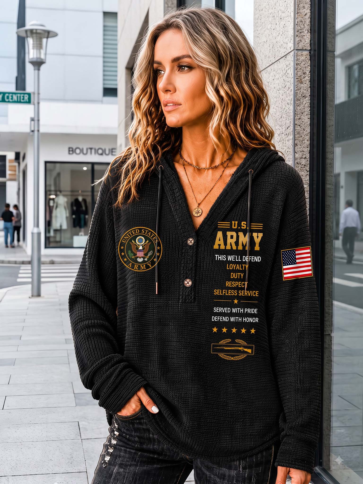 A woman wearing a black waffle-knit pullover hoodie with gold and white U.S. Army graphics, standing on a city sidewalk.