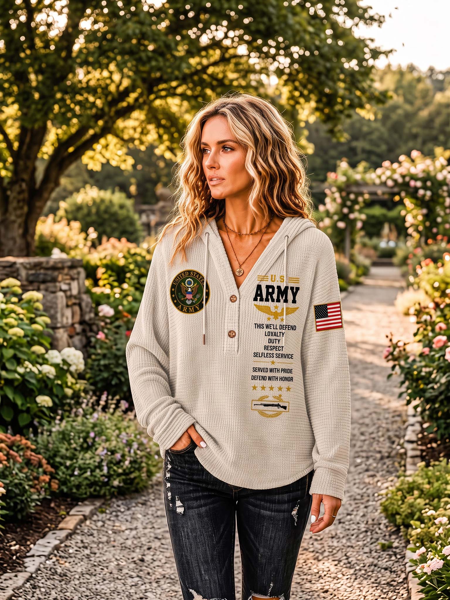 A woman stands in a sunlit garden wearing a light beige waffle-knit hoodie with military-themed graphics, including 'U.S.