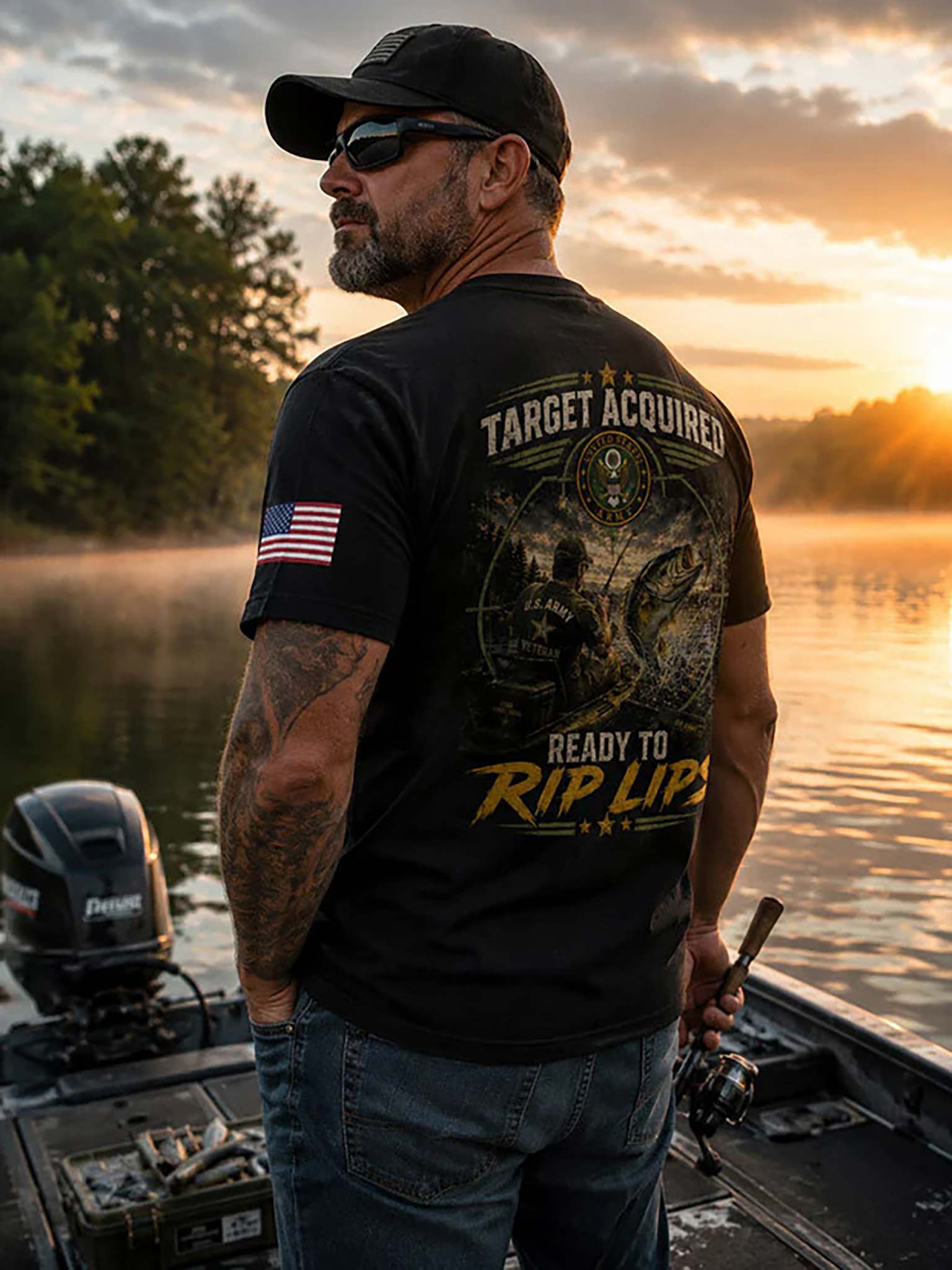 A man wearing a black graphic t-shirt with a fishing design and 'TARGET ACQUIRED READY TO RIP LIPS' text, standing on a boat on a misty lake at sunset.