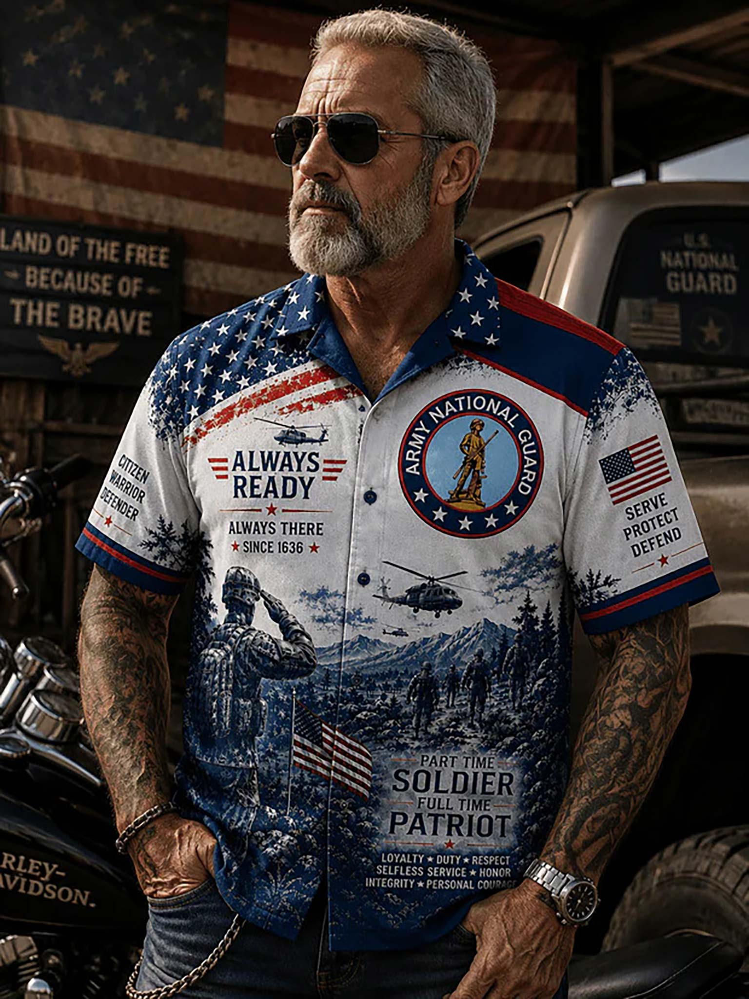 A man with a gray beard and sunglasses wearing a patriotic military-themed short-sleeve button-up shirt in a rustic garage setting with a motorcycle and truck in the background.