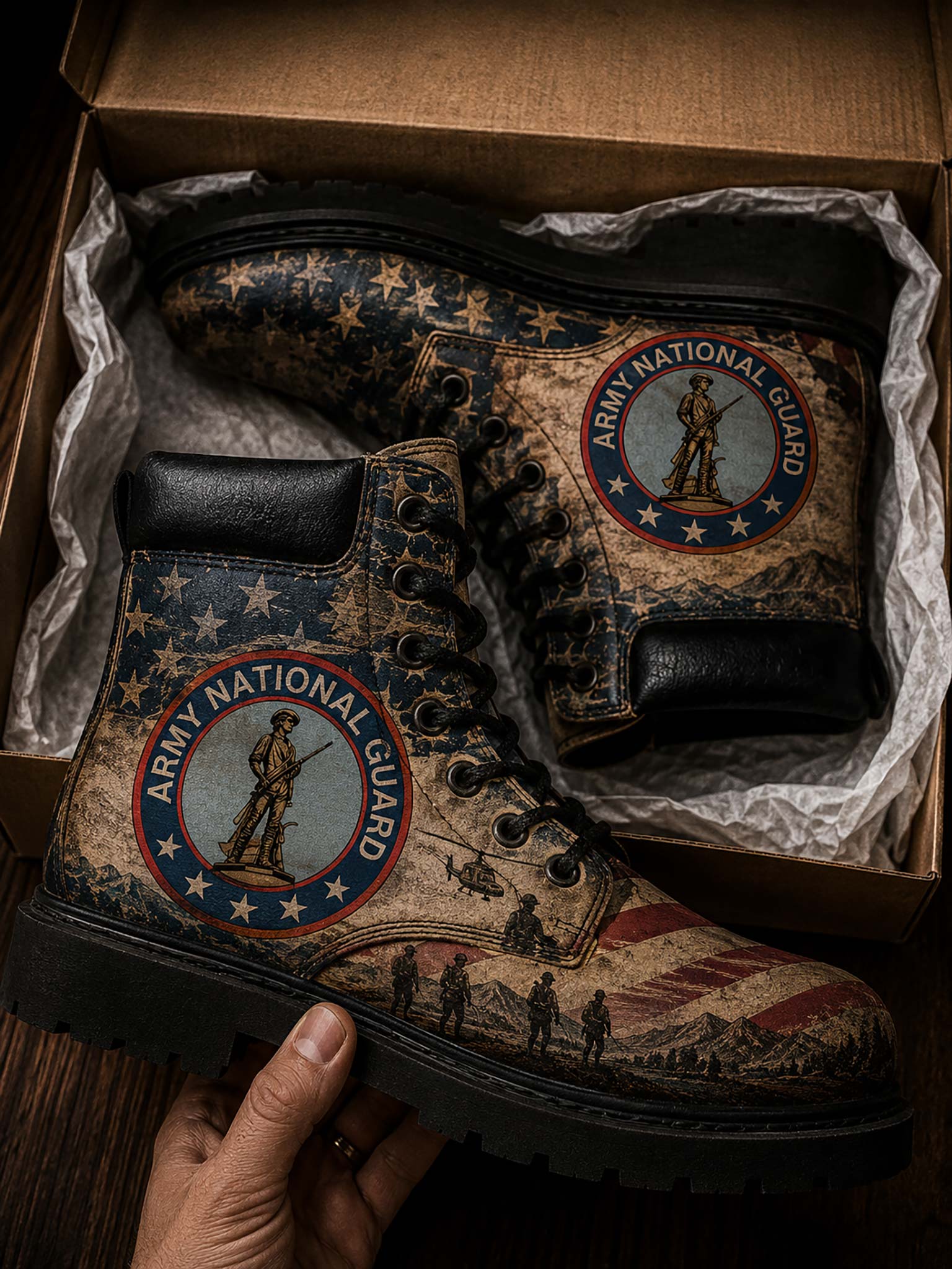 A pair of lace-up combat boots displayed inside an open cardboard shoebox with tissue paper, with a hand holding the front boot. The boots feature a distressed American flag and military theme with the 'Army National Guard' emblem on the side.