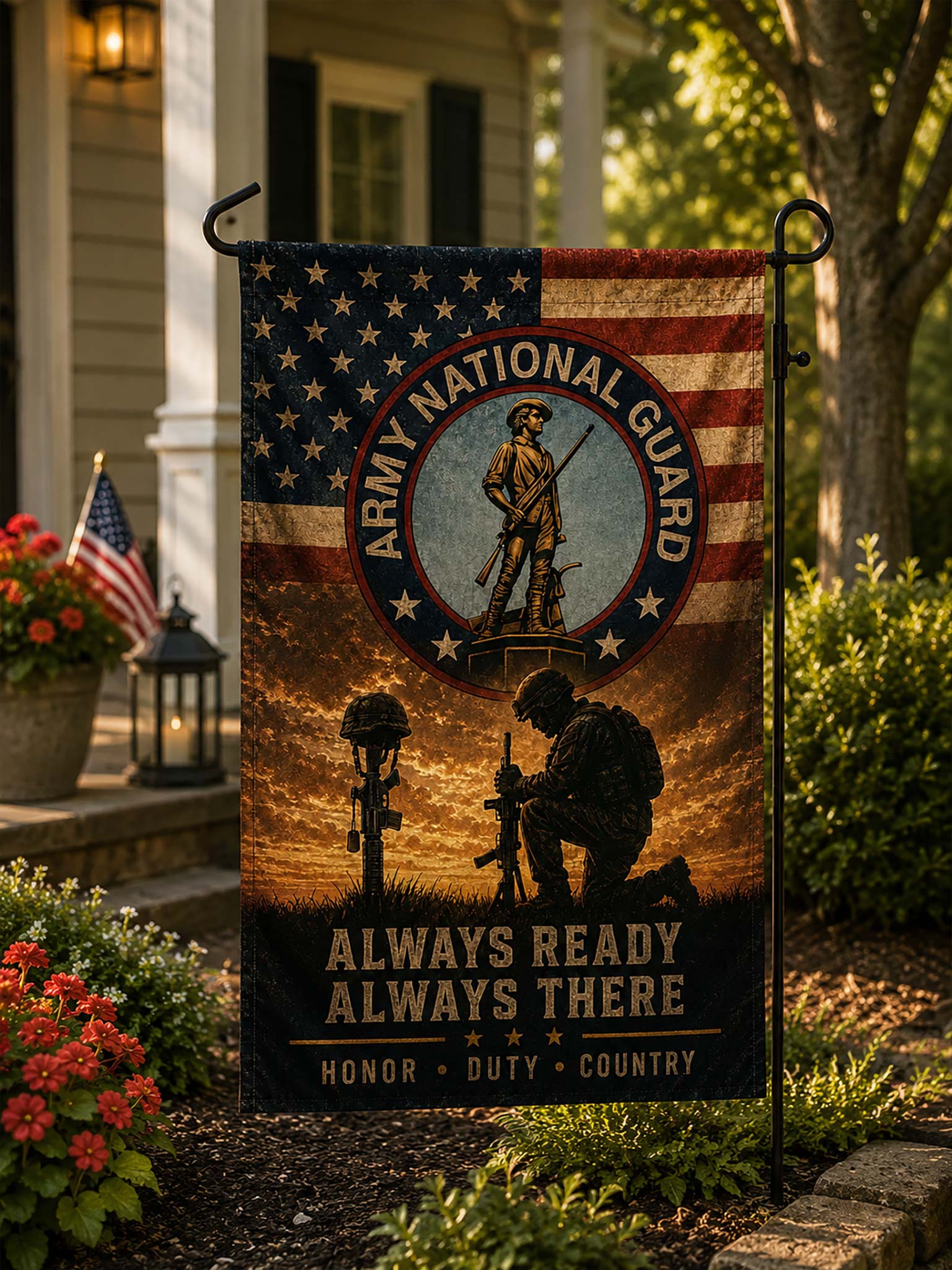 A vertical garden flag featuring the Army National Guard emblem and patriotic military imagery, displayed on a black metal stand in a landscaped garden in front of a house.