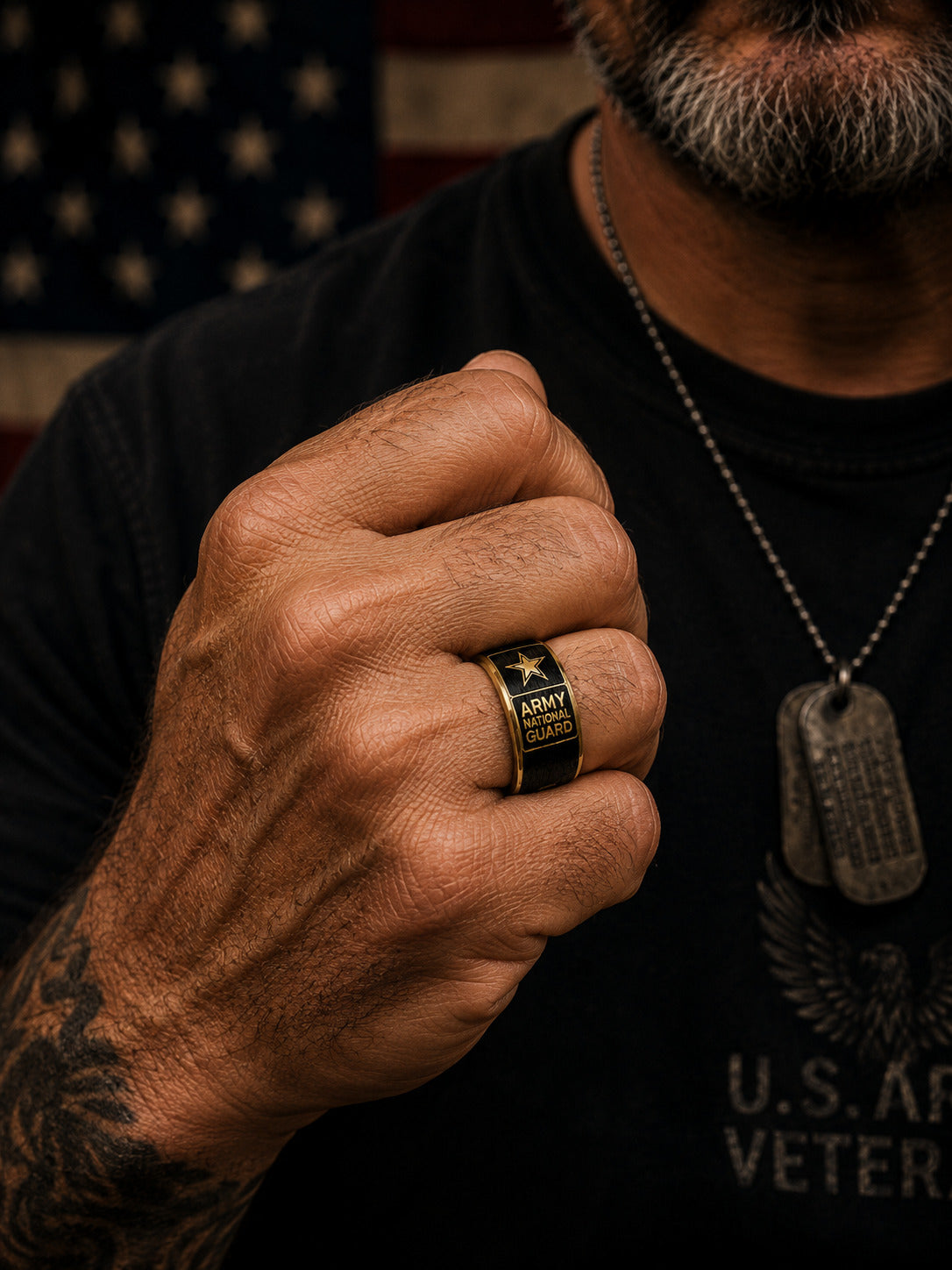 A close-up of a man's hand wearing a black and gold military-style ring engraved with 'ARMY VETERAN' and a star, while wearing a black t-shirt and dog tag necklace in front of an American flag.