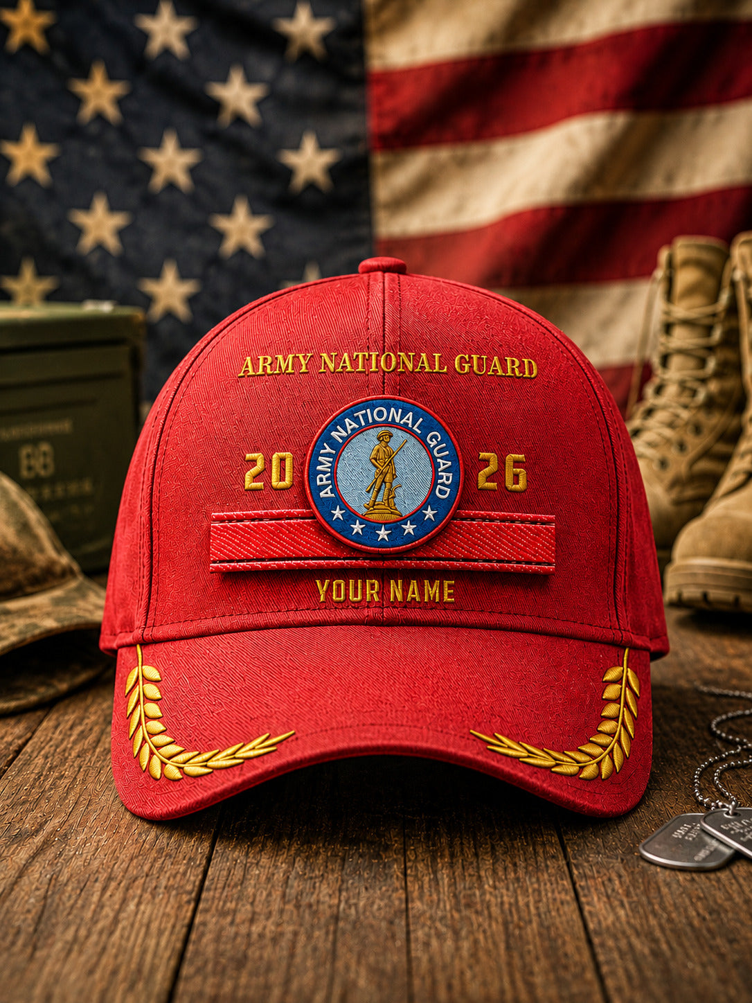 A red baseball cap with embroidered Army National Guard insignia, including a circular patch, '2026' text, and 'YOUR NAME' customization option, displayed on a wooden surface with an American flag, boots, and military accessories in the background.