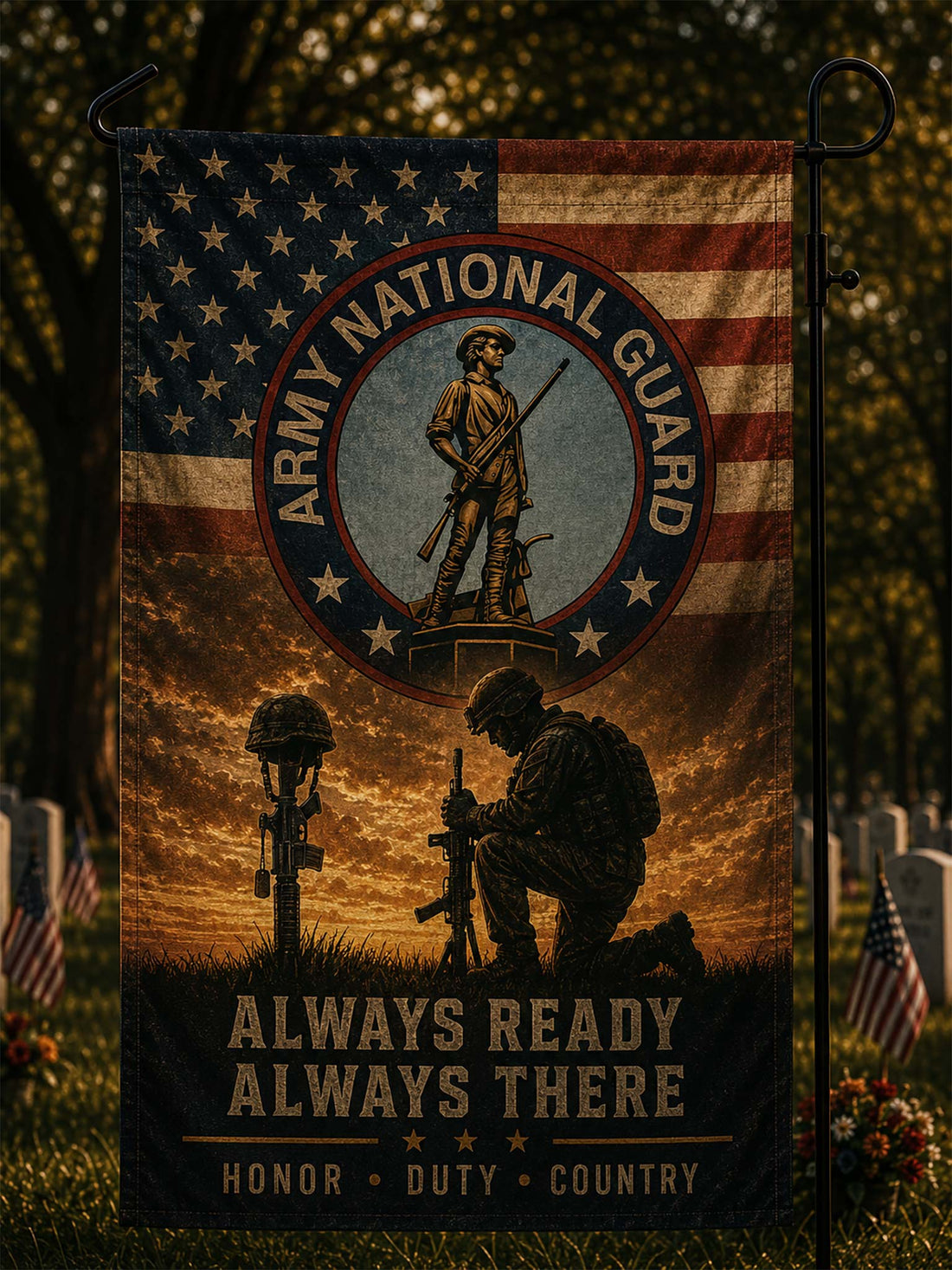 A vertical garden flag displayed on a black metal stand in an outdoor cemetery setting. The flag features a distressed American flag background with the Army National Guard emblem at the top, a kneeling soldier silhouette, a rifle on a headstone, and the text 'ALWAYS READY ALWAYS THERE' and 'HONOR • DUTY • COUNTRY'.