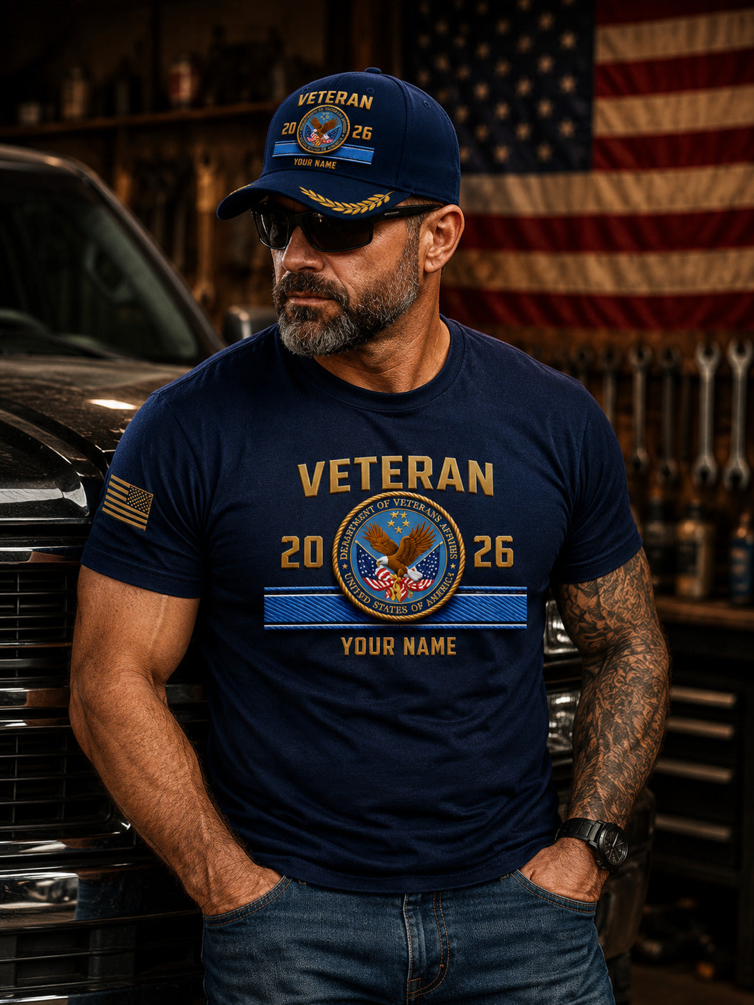 A man wearing a navy blue custom 'Veteran 2026' graphic t-shirt and matching baseball cap, standing in a garage setting with a truck and American flag in the background.