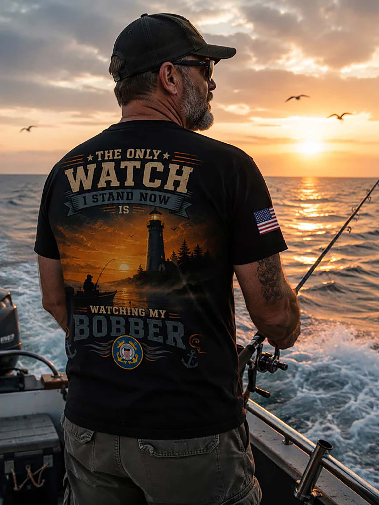 A man on a boat at sunset wearing a black graphic t-shirt with a large fishing-themed print on the back, including a lighthouse, fish, and text reading 'THE ONLY WATCH I STAND NOW IS WATCHING MY BORBER'.
