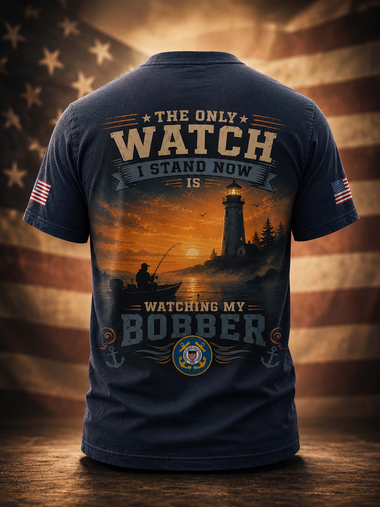 A navy blue short-sleeve t-shirt shown from the back, featuring a large graphic print of a lighthouse at sunset with a fisherman in a boat and the text 'THE ONLY WATCH I STAND NOW IS WATCHING MY BOBBER'. Small American flag graphics appear on both sleeves, and the shirt is displayed against a blurred American flag background.