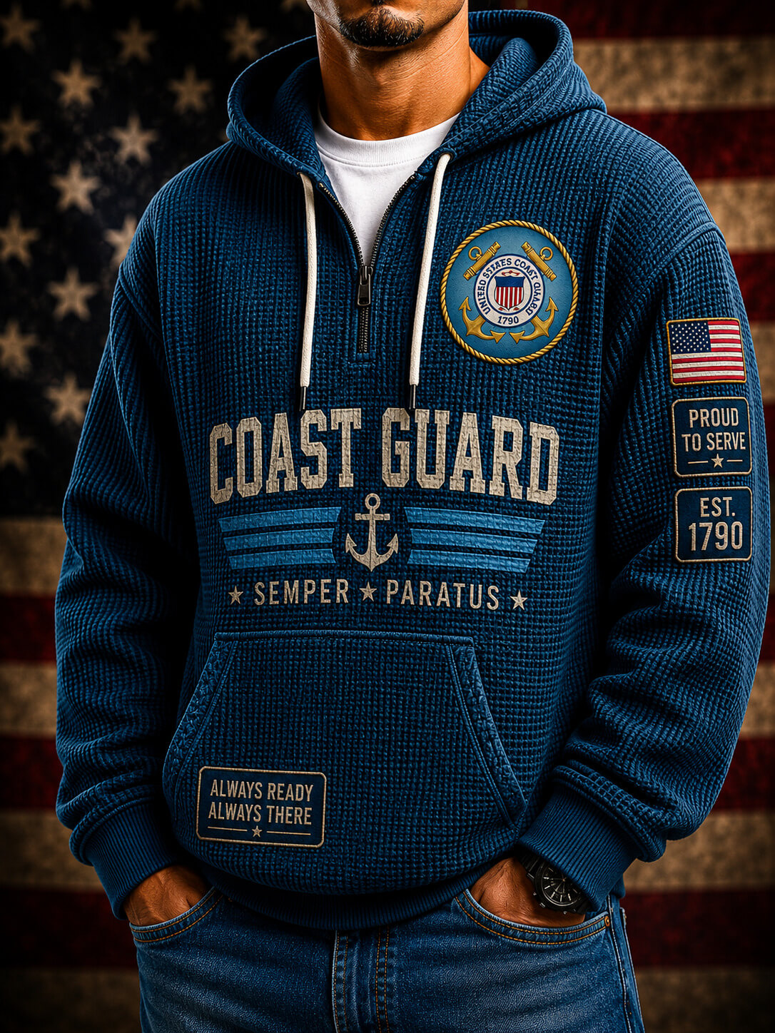 A man wearing a navy blue waffle-knit hoodie with Coast Guard graphics, including a large 'COAST GUARD' text print, an anchor emblem, and a US flag patch on the sleeve, posed against a blurred American flag background.