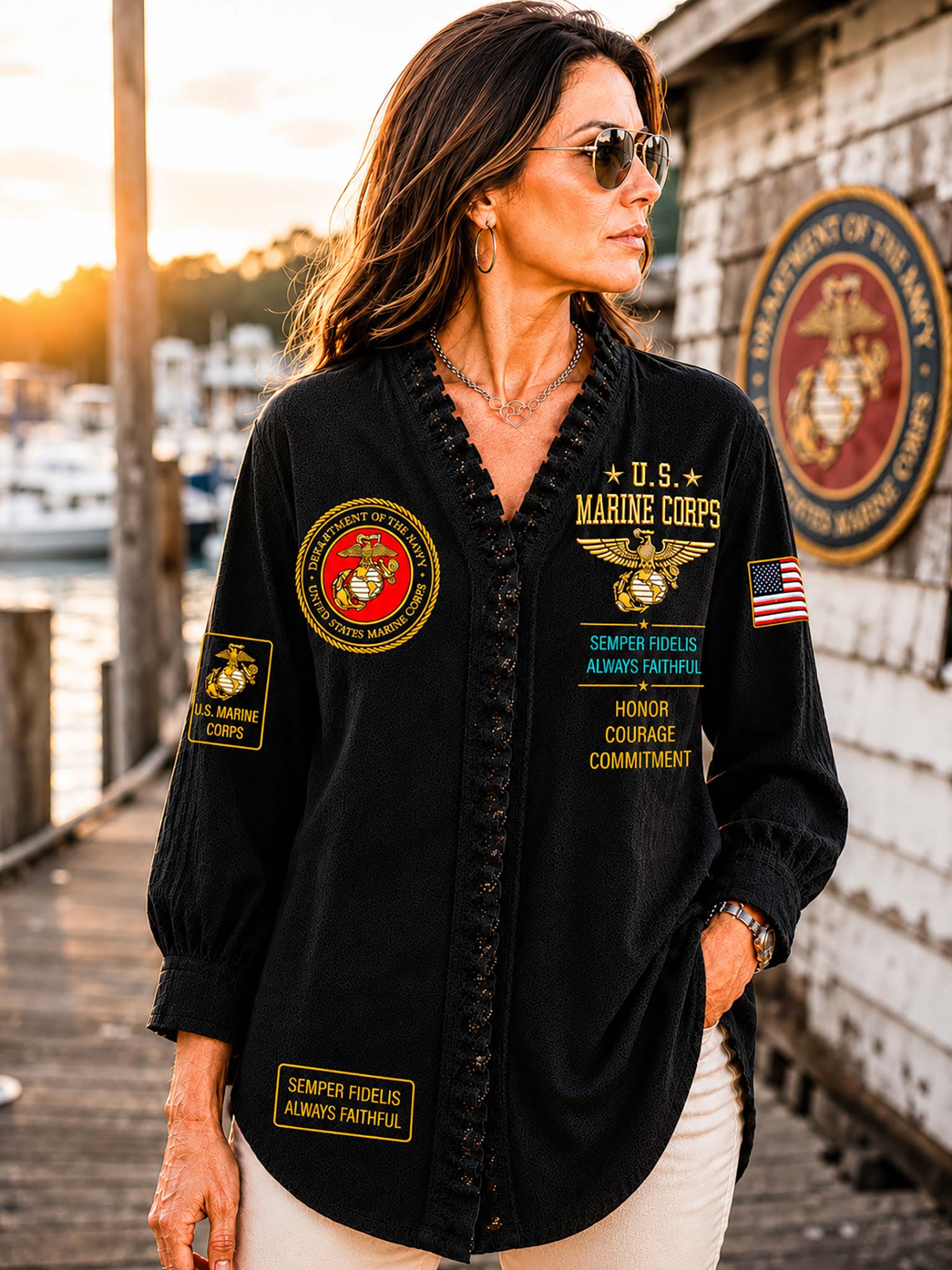 A woman wearing a black long-sleeve button-up shirt with military-themed patches and text, standing outdoors near a dock and building wall at sunset.