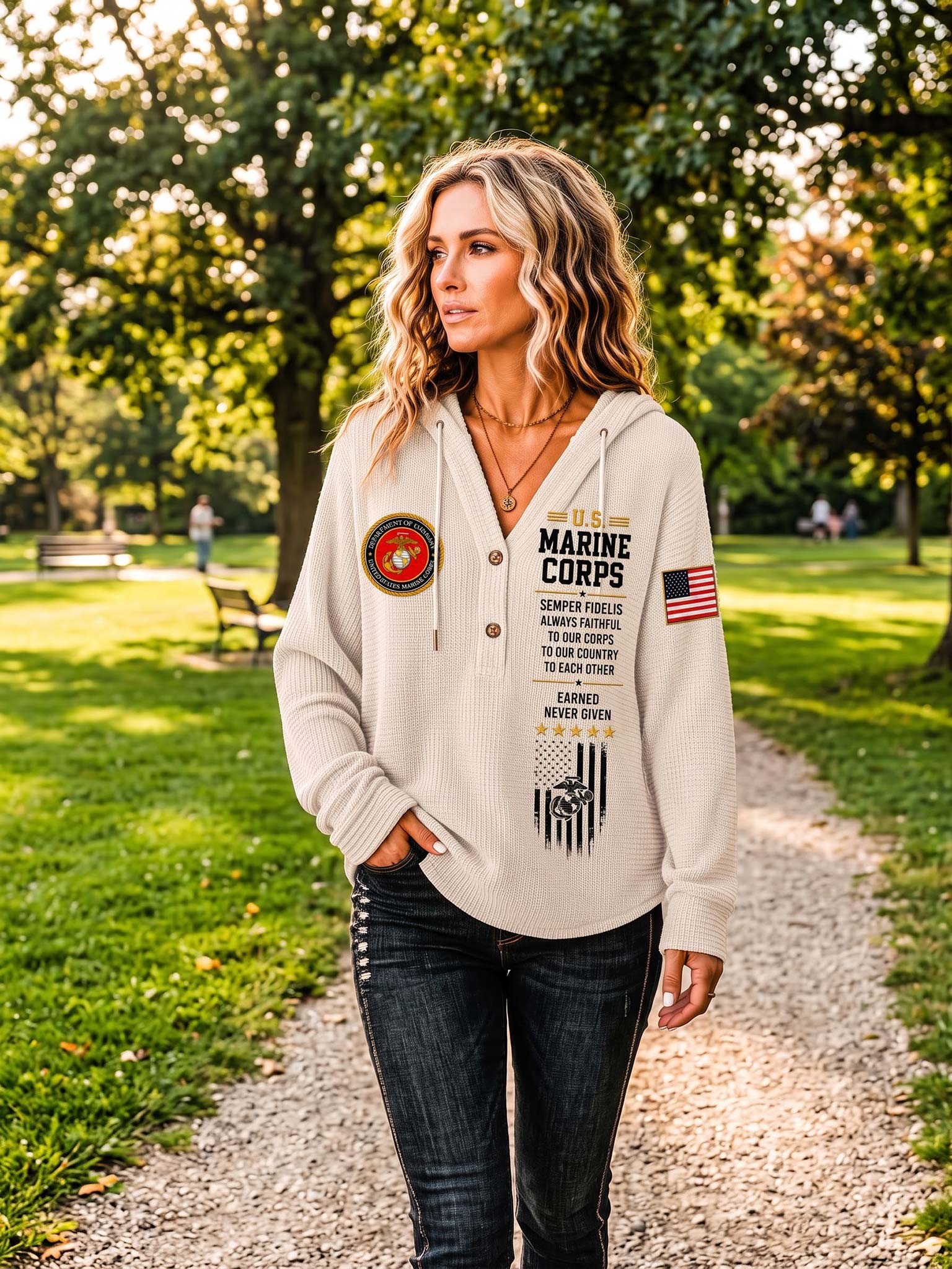 A woman stands on a gravel path in a sunny park wearing a light beige hoodie with Marine Corps graphics, including a circular emblem on the left chest, vertical text on the right chest, and an American flag patch on the sleeve, paired with dark jeans.