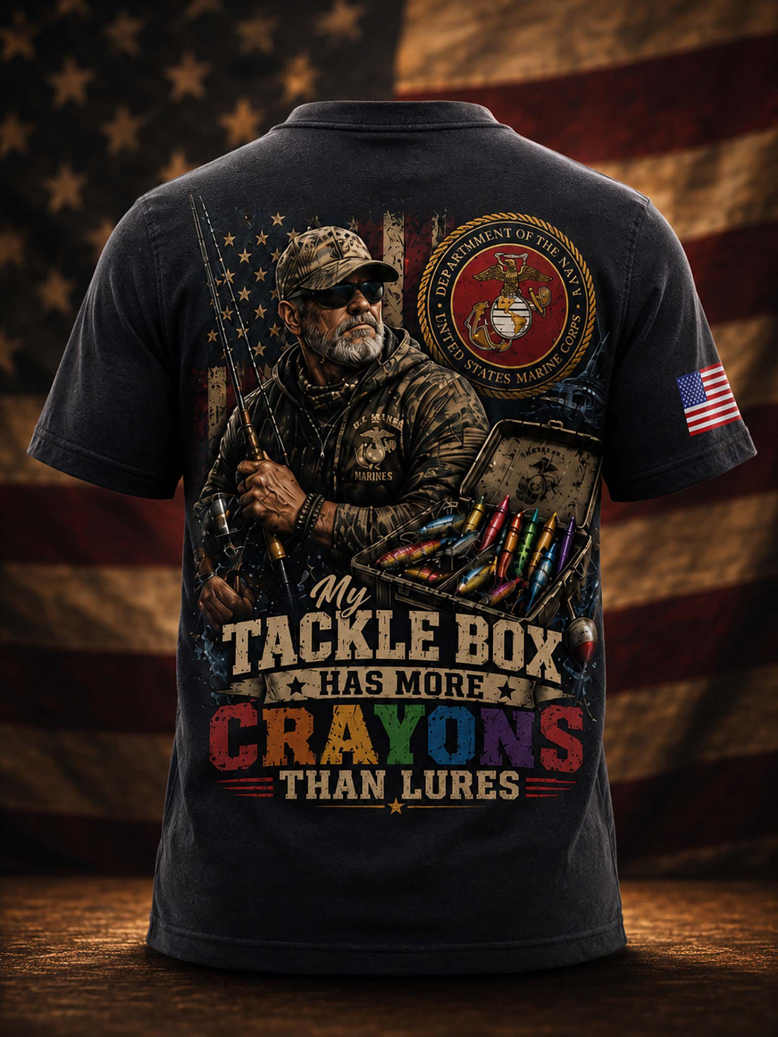 A dark navy graphic t-shirt shown from the back, featuring a large patriotic fishing design with a Marine Corps emblem and the text 'My Tackle Box HAS MORE CRAYONS THAN LURES'. The background is a blurred American flag.