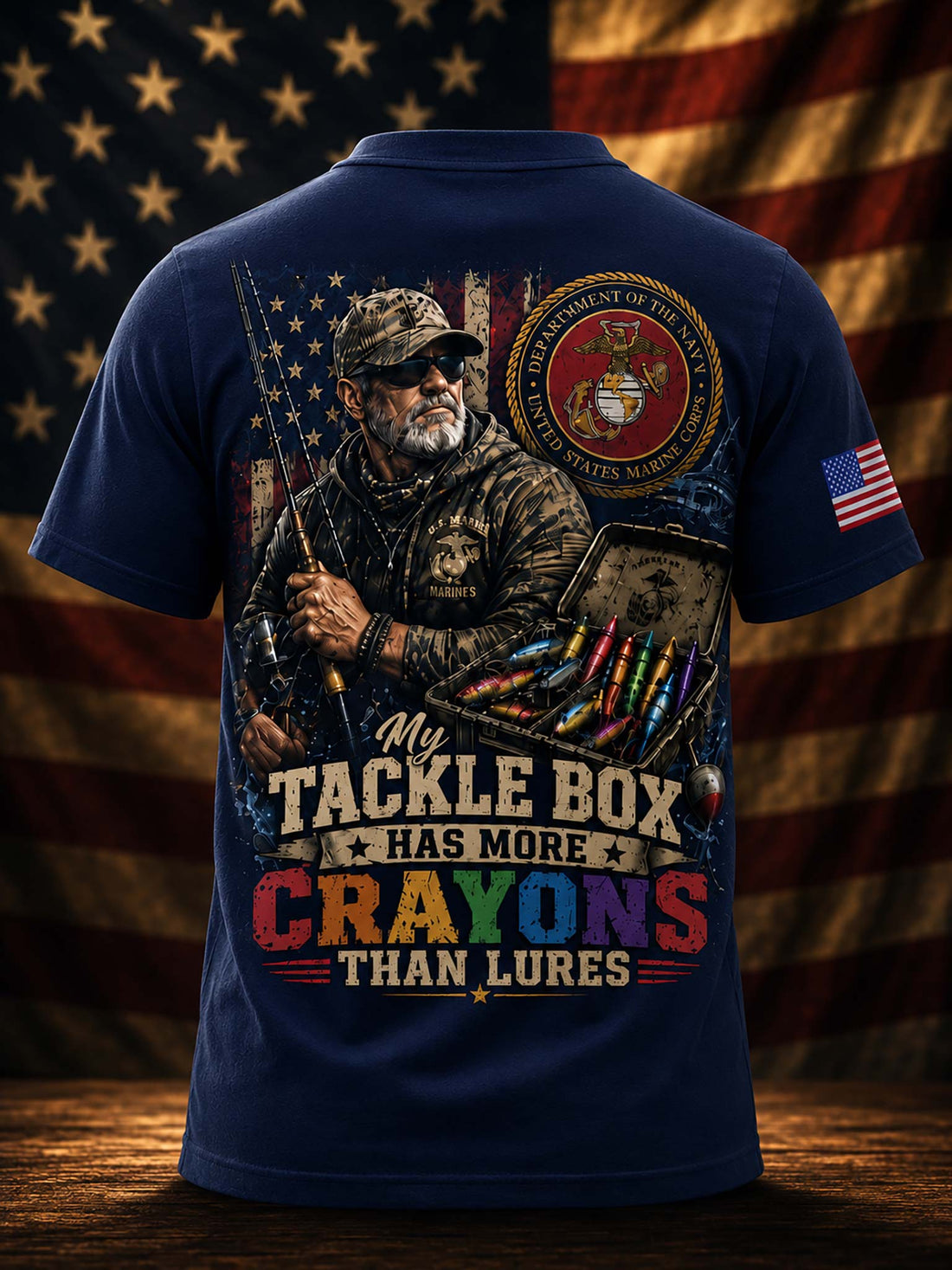A dark navy graphic t-shirt shown from the back, featuring a large patriotic fishing design with a Marine Corps emblem and the text 'My Tackle Box HAS MORE CRAYONS THAN LURES'. The background is a blurred American flag.