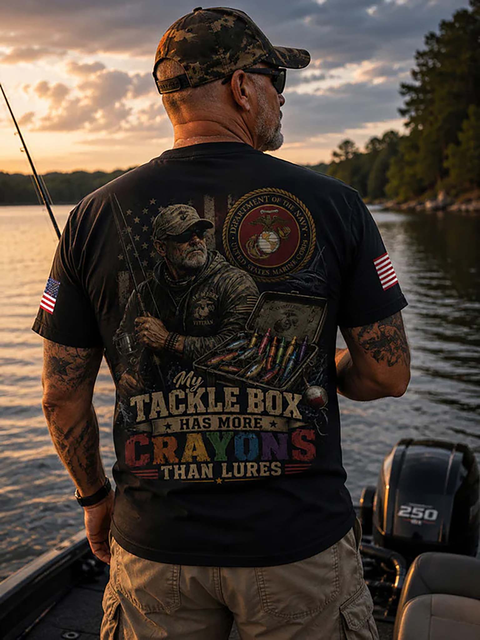 A man on a boat at sunset wearing a black graphic T-shirt with a large fishing-themed back print, khaki pants, and a camouflage cap, facing away from the camera.