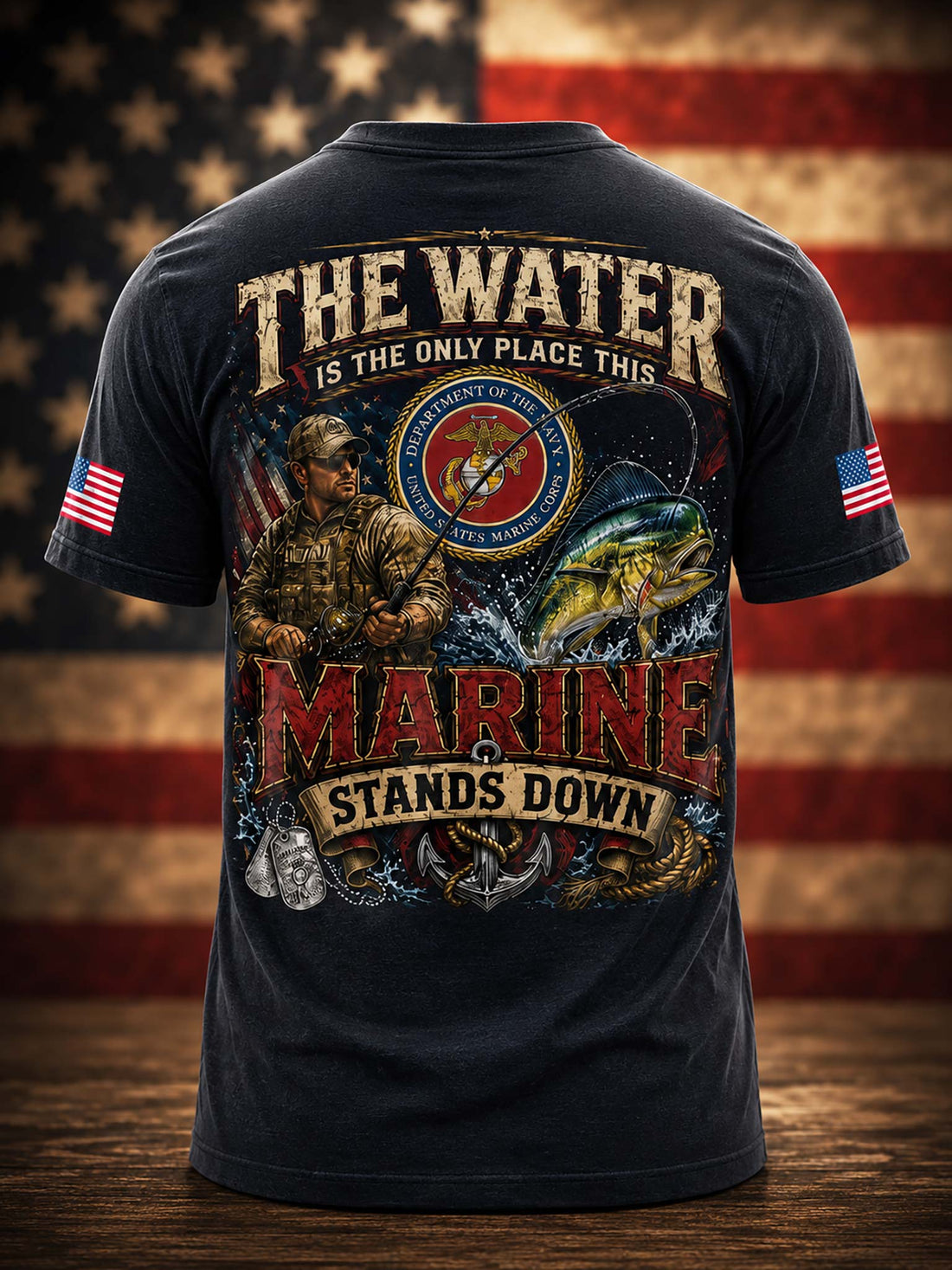 A black graphic t-shirt shown from the back, featuring a large military-themed print with the text 'THE WATER IS THE ONLY PLACE THIS MARINE STANDS DOWN' and an illustration of a Marine Corps soldier, eagle, anchor, and fish. The shirt is displayed against a blurred American flag background on a wooden surface.