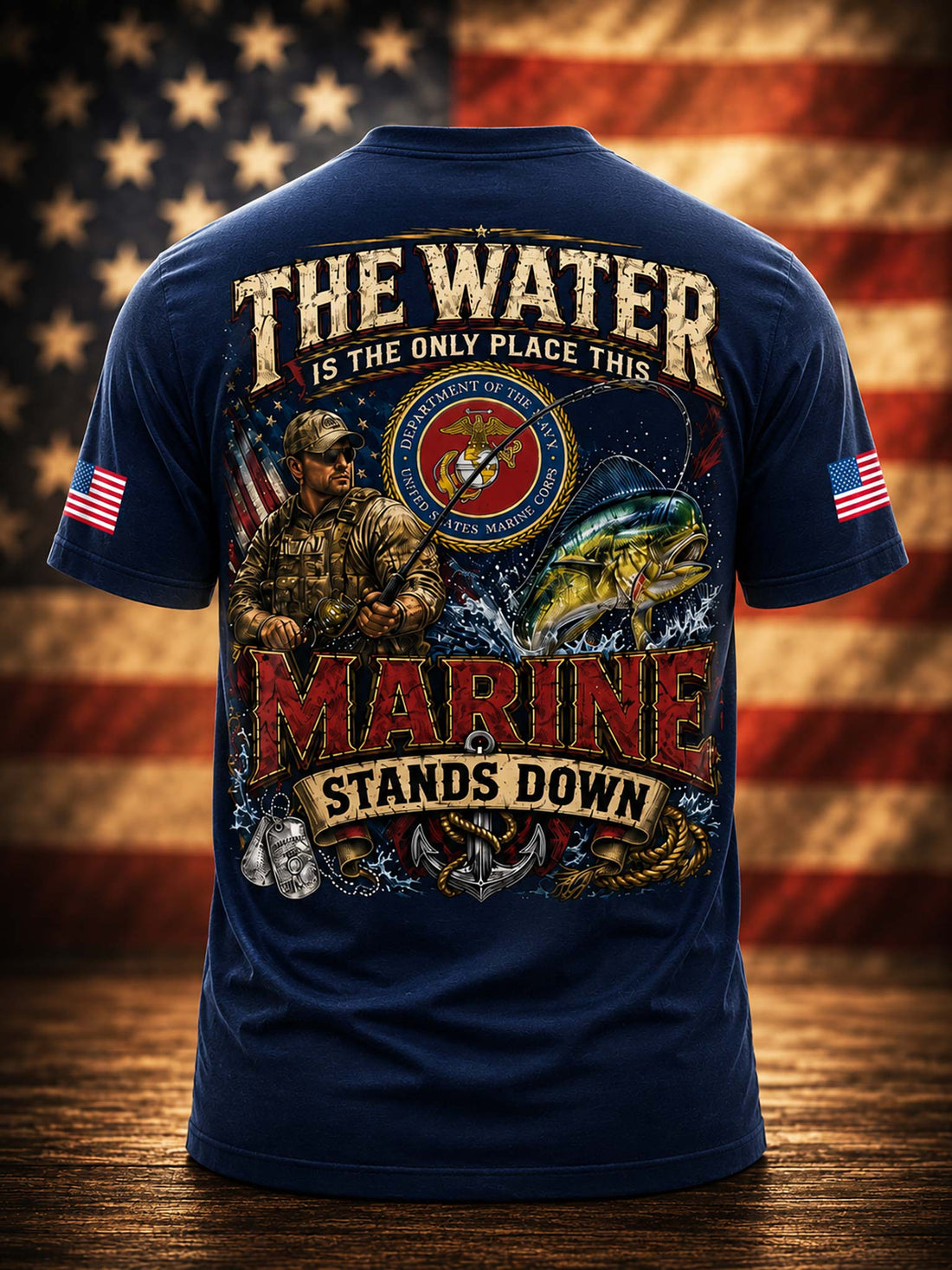 A black graphic t-shirt shown from the back, featuring a large military-themed print with the text 'THE WATER IS THE ONLY PLACE THIS MARINE STANDS DOWN' and an illustration of a Marine Corps soldier, eagle, anchor, and fish. The shirt is displayed against a blurred American flag background on a wooden surface.