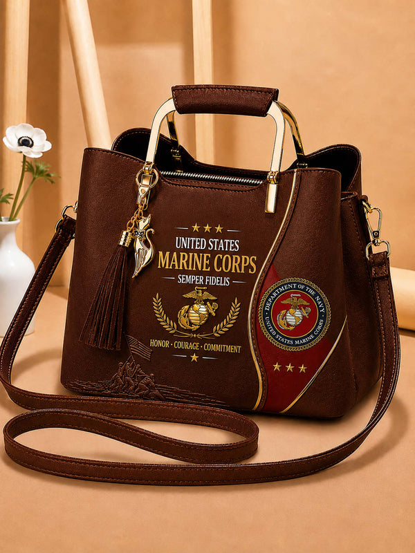A dark brown faux-leather handbag with a top handle and shoulder strap, decorated with United States Marine Corps emblems, text, and a tassel charm, set against a warm beige background.