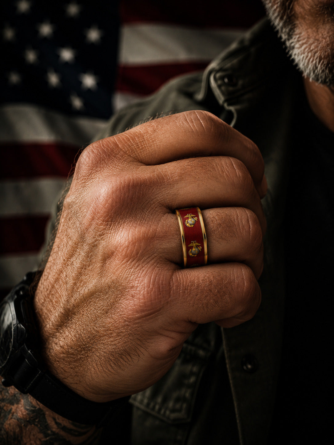 A close-up of a man's clenched fist wearing a gold ring with red enamel and the Eagle, Globe, and Anchor insignia, with an American flag blurred in the background.