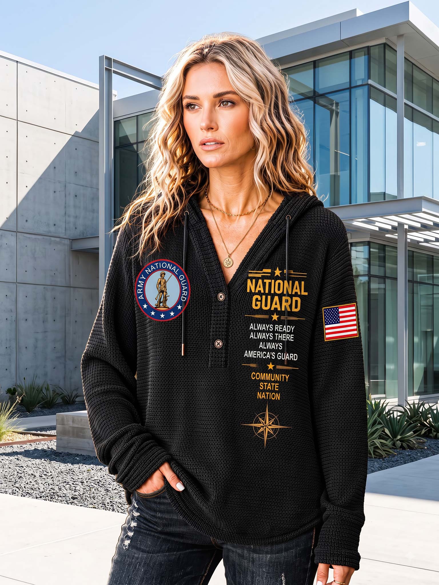 A woman wearing a black waffle-knit pullover hoodie with 'National Guard' graphics, standing outdoors in front of a modern glass building.