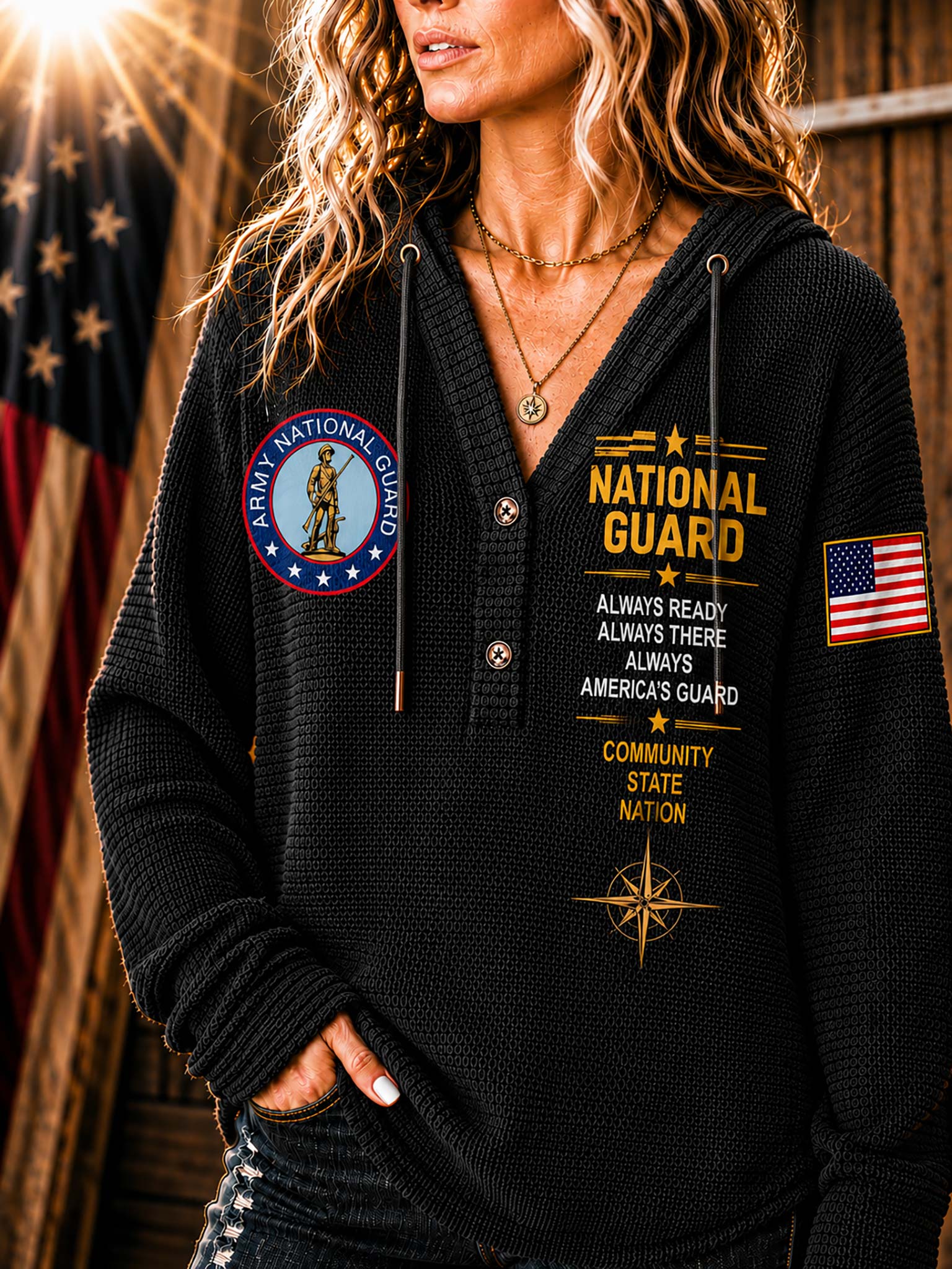 A woman wearing a black waffle-knit hoodie with 'National Guard' text and graphics on the front and sleeves, photographed in a rustic indoor setting with an American flag in the background.
