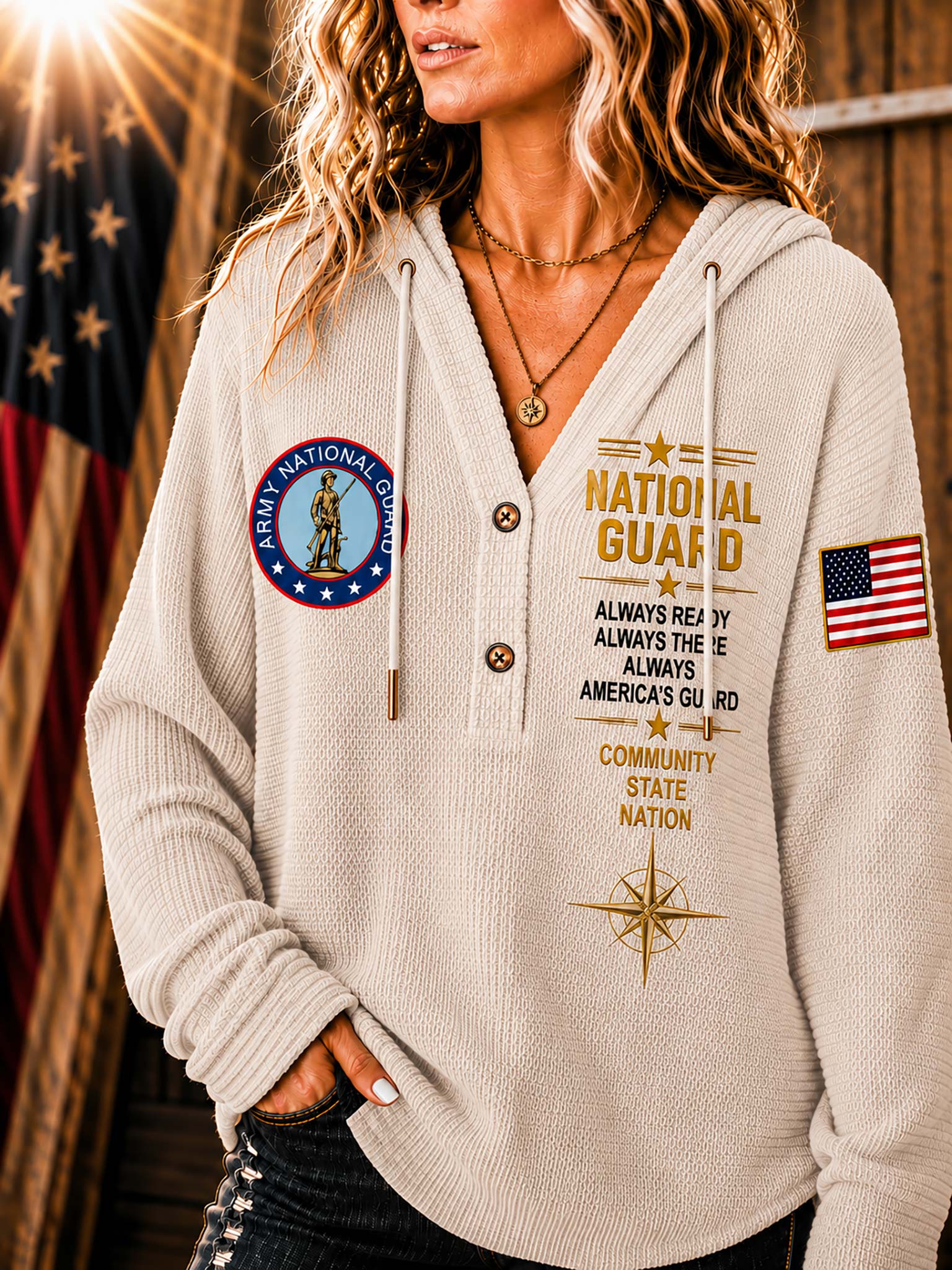 A woman wearing a cream-colored waffle-knit hoodie with 'National Guard' graphics, including a circular emblem on the left chest, gold text on the right chest, and an American flag patch on the sleeve, set against a rustic background with a blurred American flag.