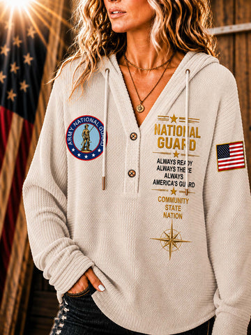 A woman wearing a cream-colored waffle-knit hoodie with 'National Guard' graphics, including a circular emblem on the left chest, gold text on the right chest, and an American flag patch on the sleeve, set against a rustic background with a blurred American flag.