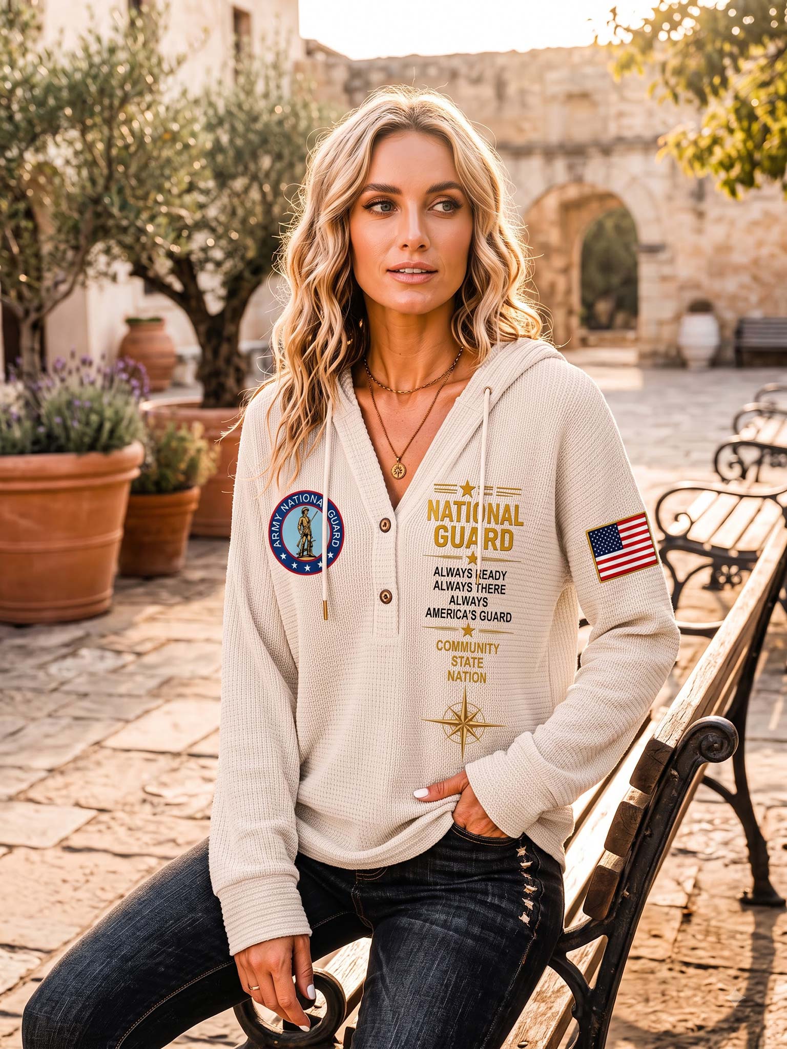 A woman sitting on a park bench wearing a cream-colored waffle-knit hoodie with 'National Guard' graphics, in an outdoor stone courtyard setting.