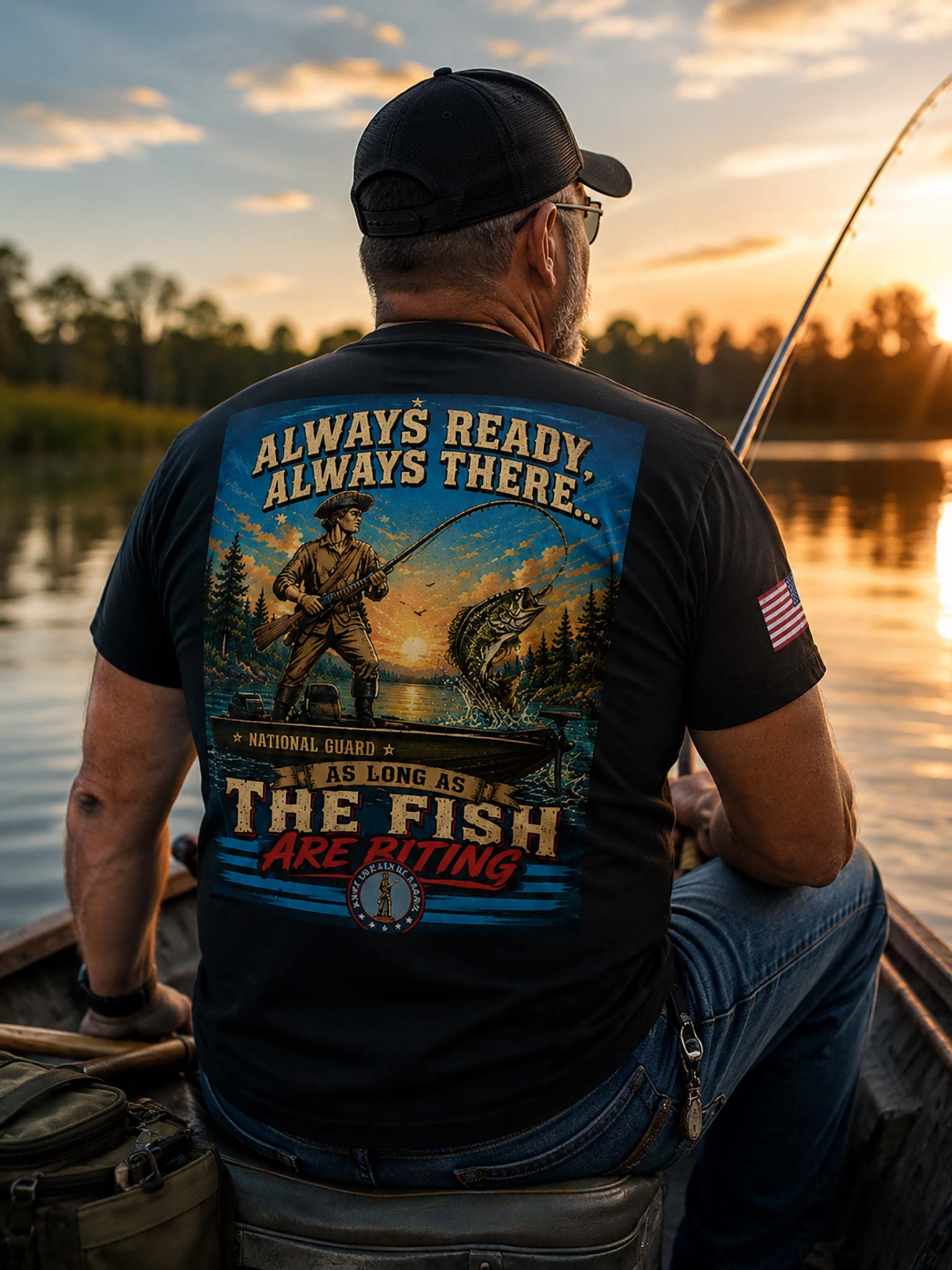 A man wearing a black t-shirt with a large back graphic and a black cap, sitting in a boat on a lake at sunset. The graphic features a fisherman, a large fish, and the text 'ALWAYS READY ALWAYS THERE.