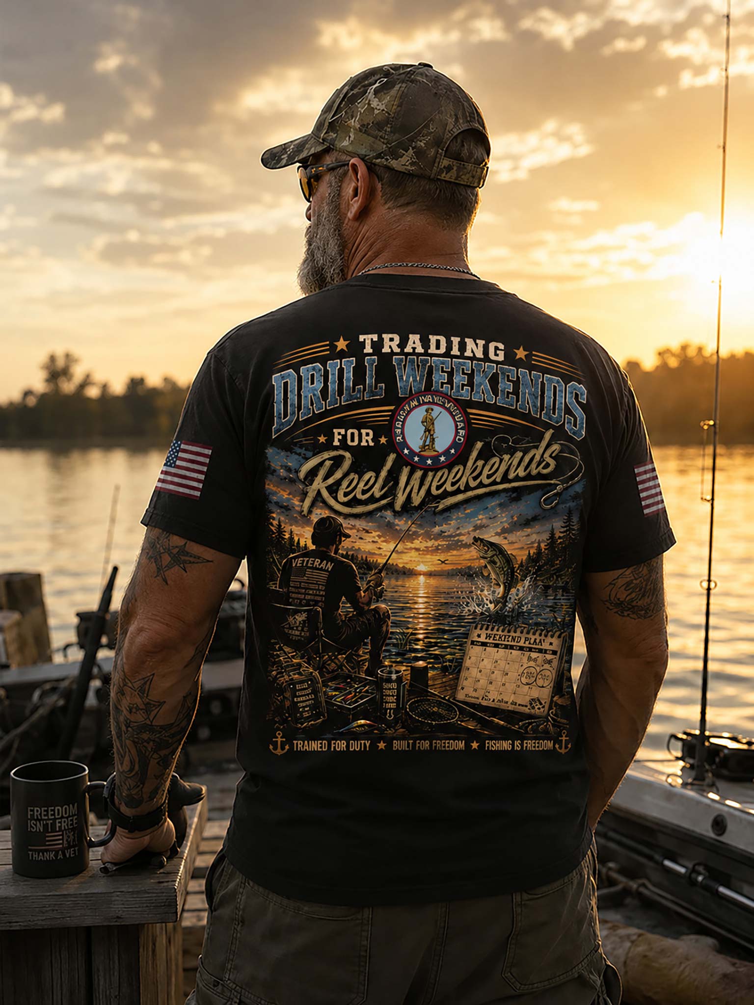 A man stands on a dock at sunset wearing a black graphic T-shirt with a large back print reading 'Trading Drill Weekends for Reel weekends' and a fishing scene, plus camouflage cap and sunglasses.
