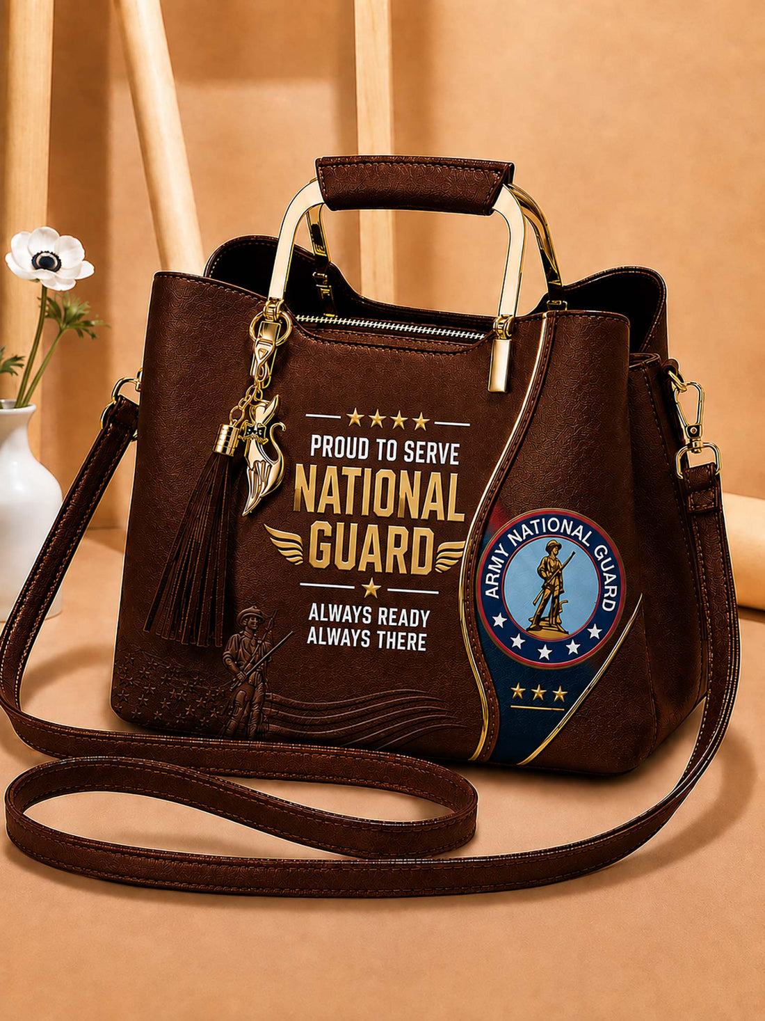 A dark brown faux-leather handbag with a top handle and shoulder strap, featuring a 'National Guard' graphic print and an Army National Guard emblem on the front, set against a warm beige background.