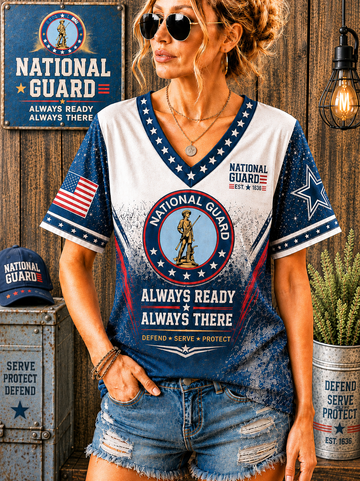 A woman wearing a distressed National Guard themed V-neck t-shirt with patriotic blue, white, and red graphics, paired with denim shorts, posed against a rustic wooden wall backdrop with themed props.