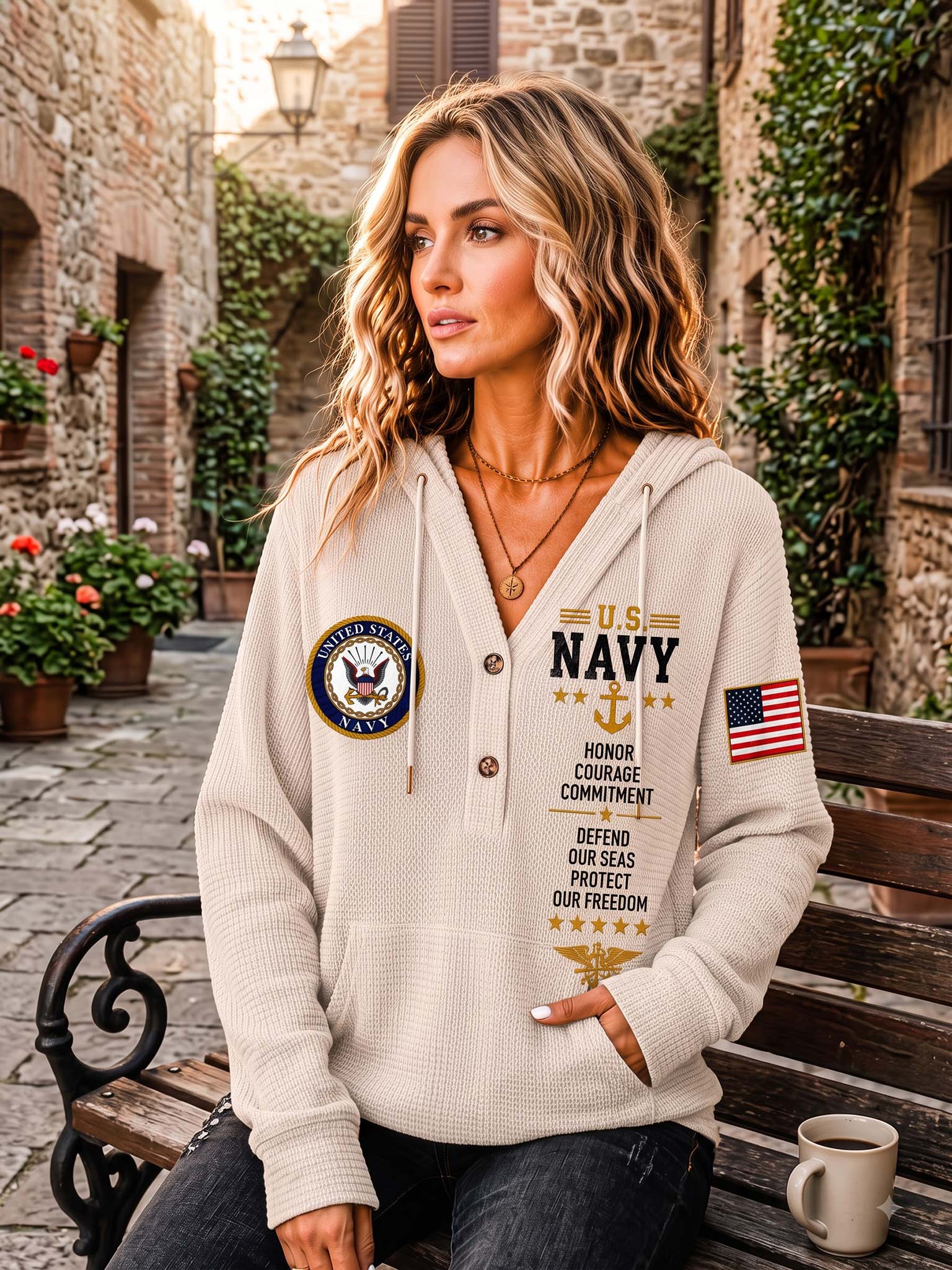 A woman sits on a wooden bench in an outdoor stone alleyway, wearing a cream-colored waffle-knit hoodie with U.S.