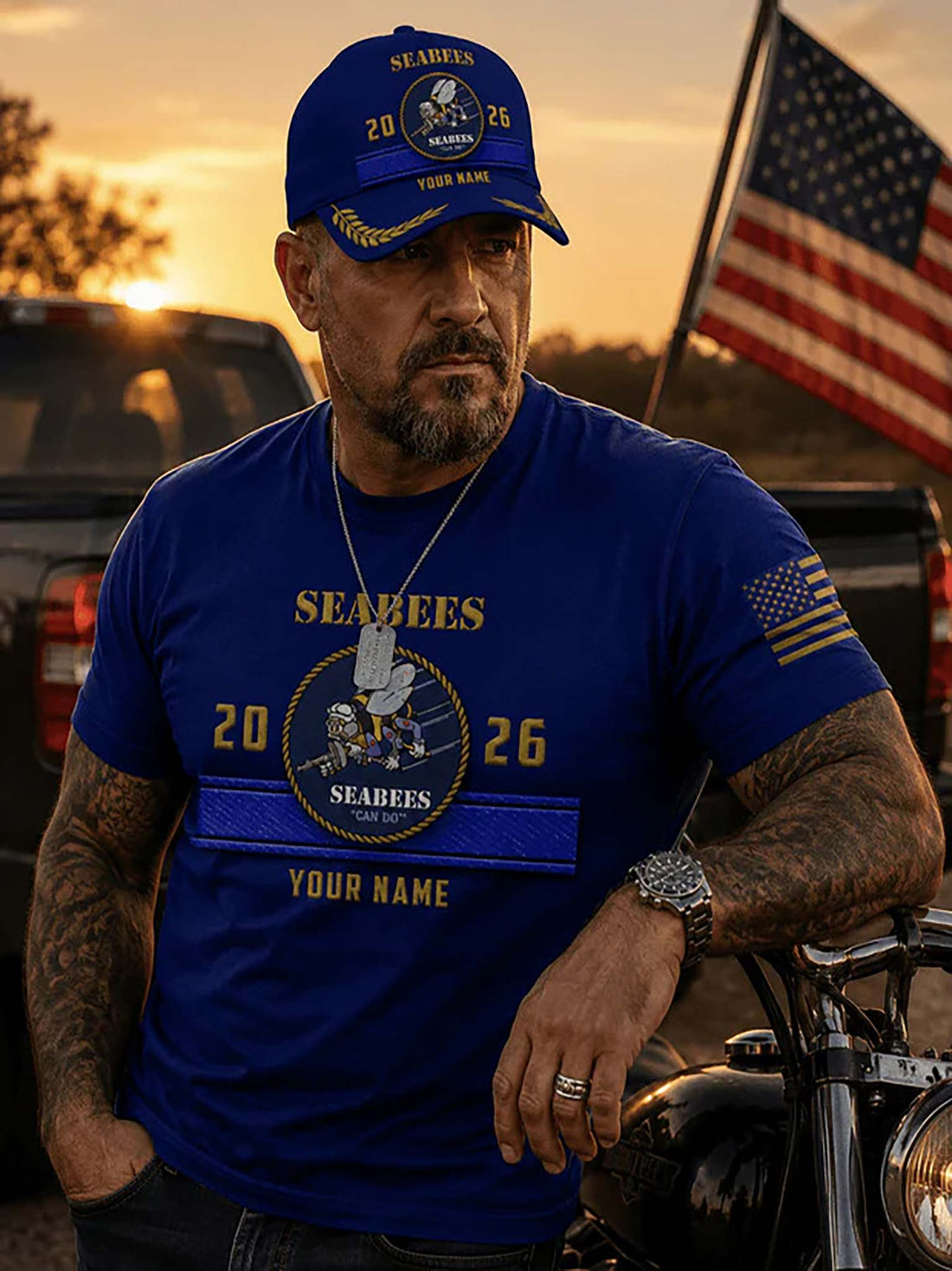 A royal blue baseball cap with gold and white embroidered Seabees insignia, featuring 'SEABEES', '2026', 'YOUR NAME', and laurel wreath embroidery on the brim, set against a rustic wooden surface with a blurred American flag background.