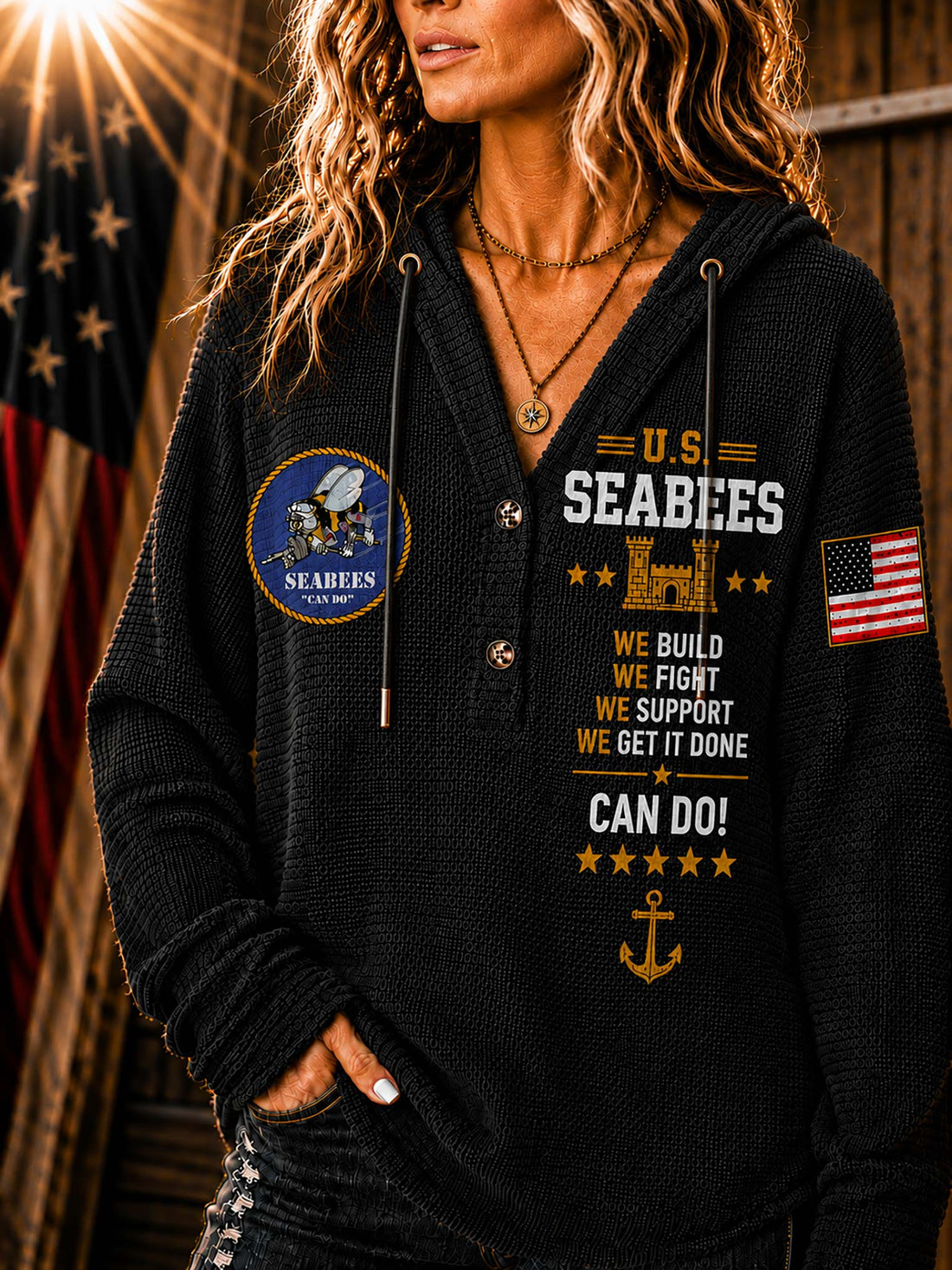 A woman wearing a cream-colored ribbed hoodie with US Seabees graphics, including a circular emblem on the left chest, text on the right chest, and an American flag patch on the sleeve, set against a rustic wooden background with a blurred American flag.