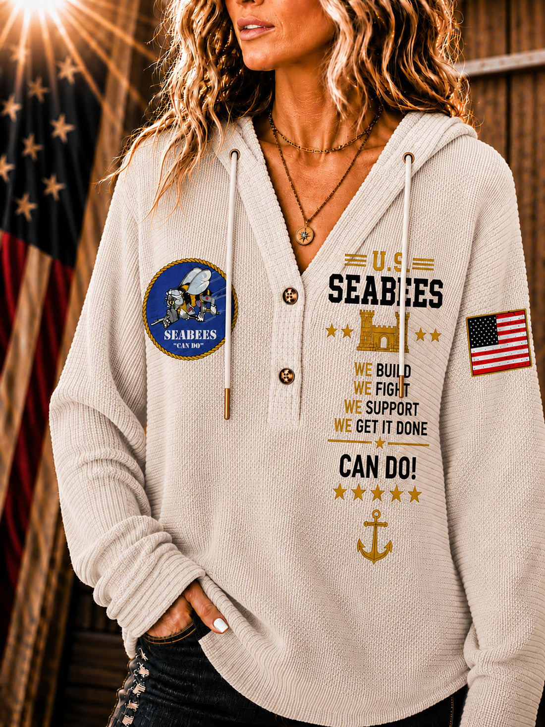 A woman wearing a cream-colored ribbed hoodie with US Seabees graphics, including a circular emblem on the left chest, text on the right chest, and an American flag patch on the sleeve, set against a rustic wooden background with a blurred American flag.