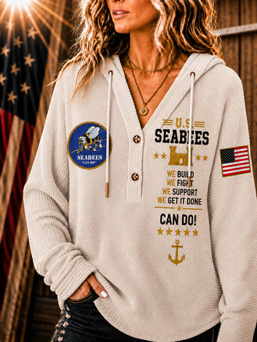 A woman wearing a cream-colored ribbed hoodie with US Seabees graphics, including a circular emblem on the left chest, text on the right chest, and an American flag patch on the sleeve, set against a rustic wooden background with a blurred American flag.