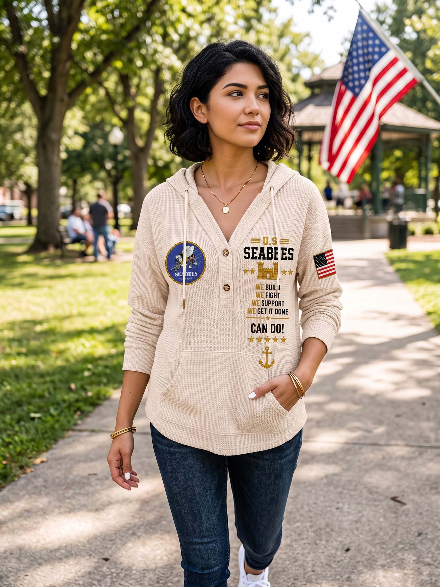 A woman wearing a beige pullover hoodie with a nautical-themed graphic print stands on a paved path in a park. The hoodie features a blue US Navy emblem on the left chest, a vertical text print reading 'SEABEES' with supporting text, a gold anchor graphic, and a small American flag on the right chest.