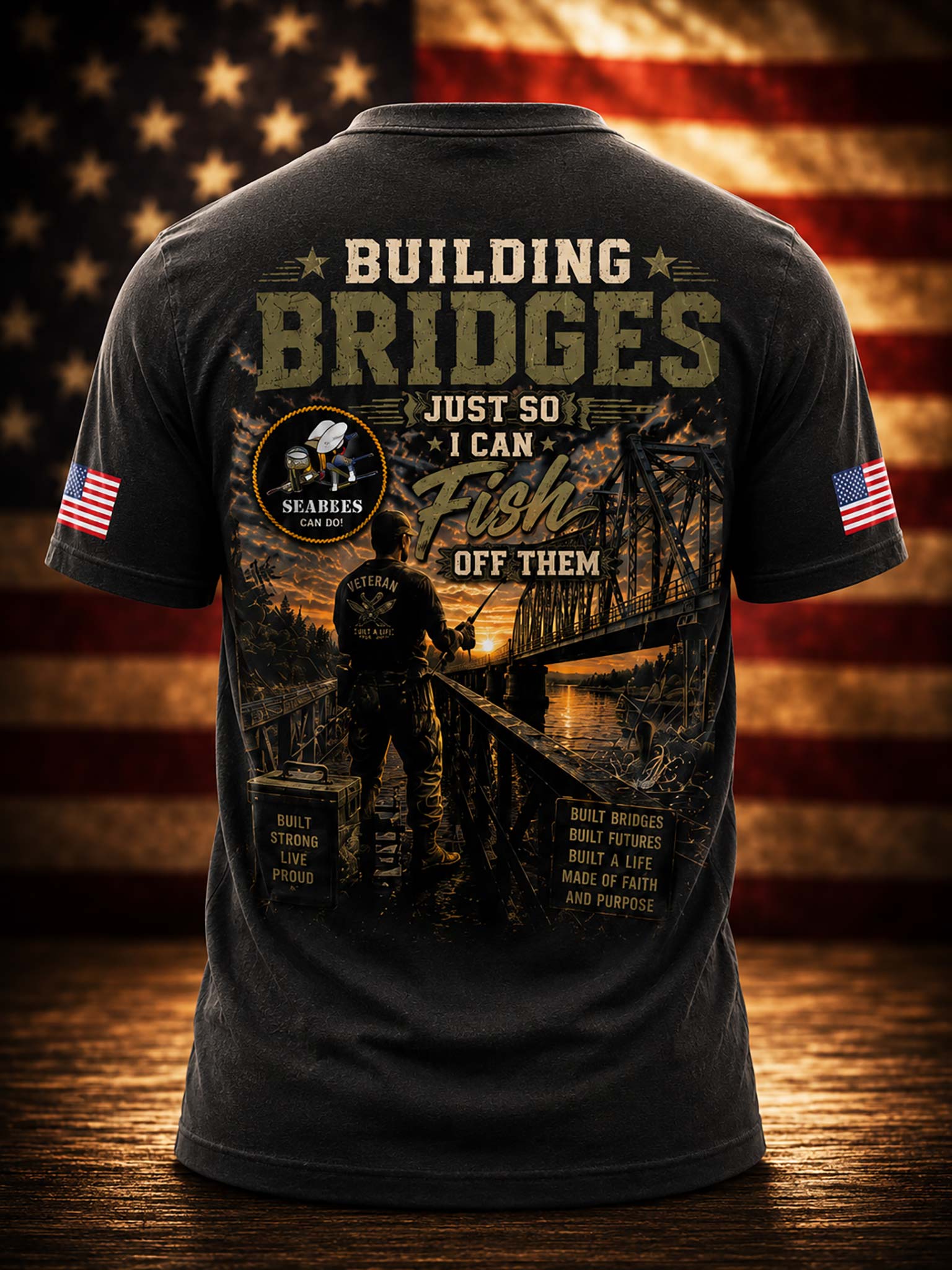 A dark charcoal gray t-shirt shown from the back, featuring a large graphic print with the text 'BUILDING BRIDGES JUST SO I CAN Fish OFF THEM' and a fisherman silhouette over a bridge scene. The shirt has American flag prints on both sleeves and is displayed against a rustic wooden background with a blurred American flag.