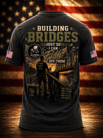 A dark charcoal gray t-shirt shown from the back, featuring a large graphic print with the text 'BUILDING BRIDGES JUST SO I CAN Fish OFF THEM' and a fisherman silhouette over a bridge scene. The shirt has American flag prints on both sleeves and is displayed against a rustic wooden background with a blurred American flag.