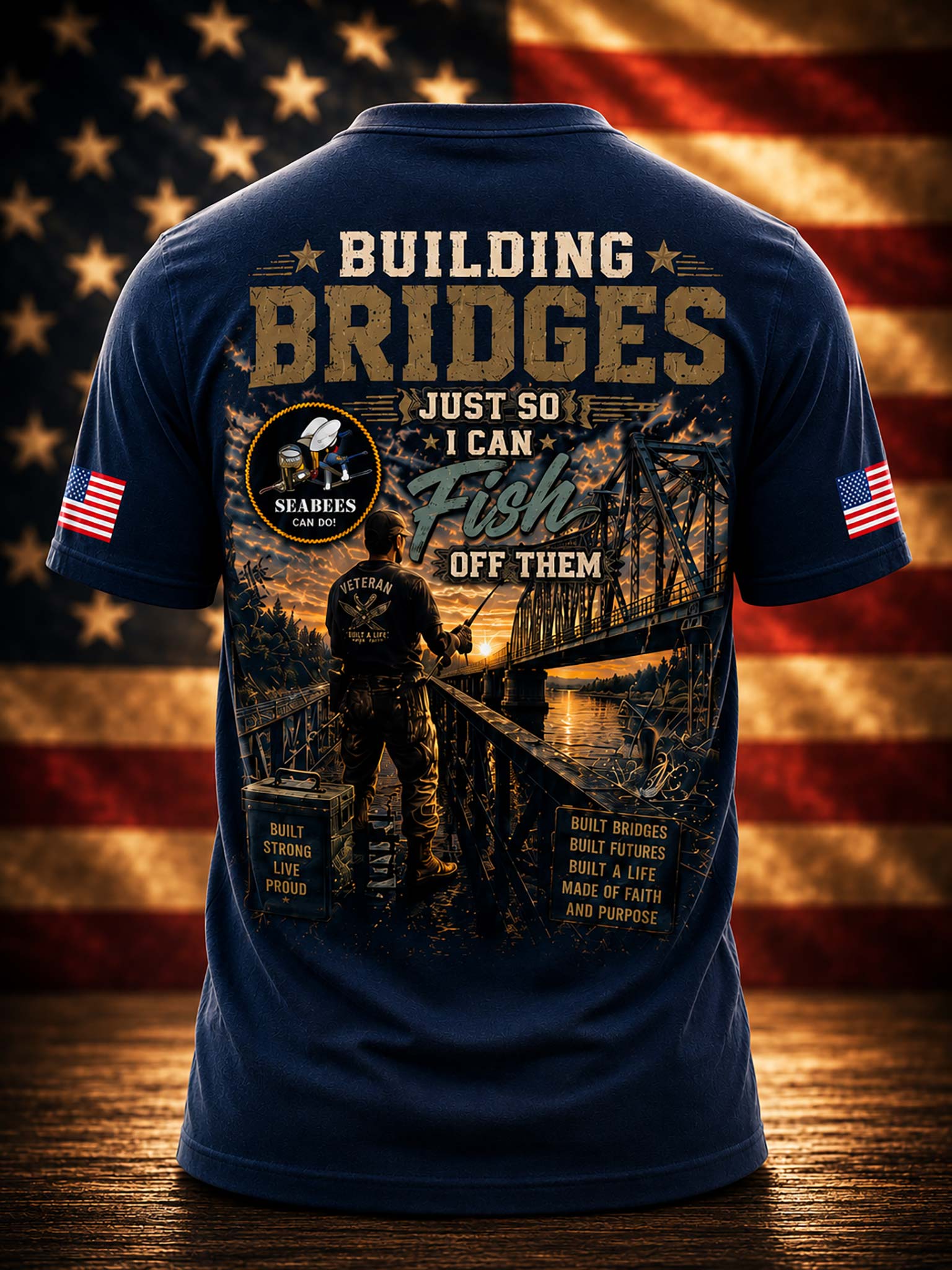 A navy blue short-sleeve t-shirt shown from the back, featuring a large patriotic fishing graphic with a bridge, a fisherman silhouette, and text including 'BUILDING BRIDGES JUST SO! I CAN Fish OFF THEM'. The shirt has small American flag prints on both sleeves and a circular 'Seabees Can Do' emblem on the upper left back.